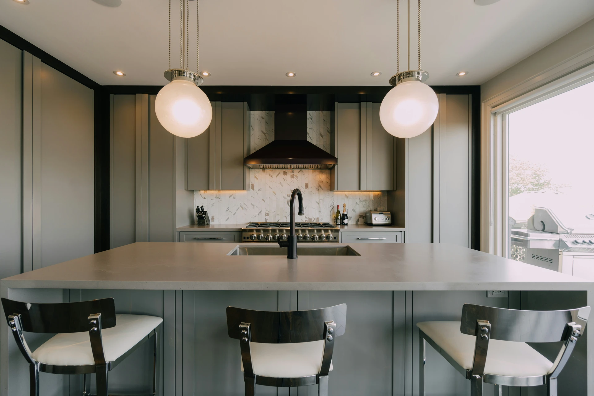 Sleek and modern kitchen featuring a spacious island with clean lines and integrated sink, embodying a minimalistic style. The muted gray tones, paired with globe pendant lights and chrome-accented stools, create a sophisticated yet understated look. Ideal for those seeking kitchen island decor ideas that emphasize simplicity, function, and elegance in a minimalistic style.