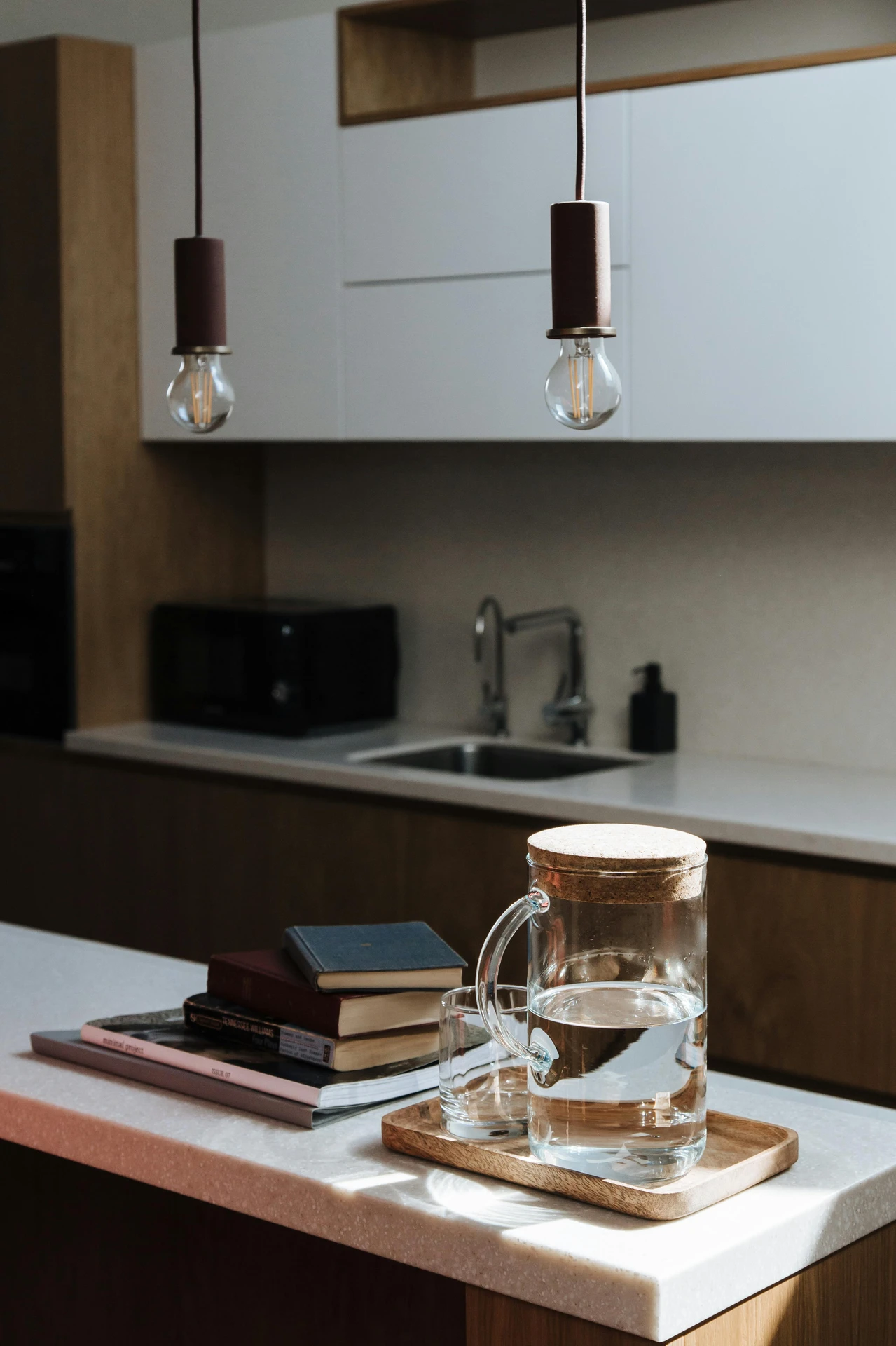 Minimalist kitchen with a clean, modern design featuring a compact island decorated with a stack of books and a wooden tray holding a glass pitcher and cup. This setup showcases thoughtful kitchen island decor ideas that incorporate personal items, creating a cozy and lived-in atmosphere. Warm lighting and natural materials enhance the inviting, understated aesthetic.