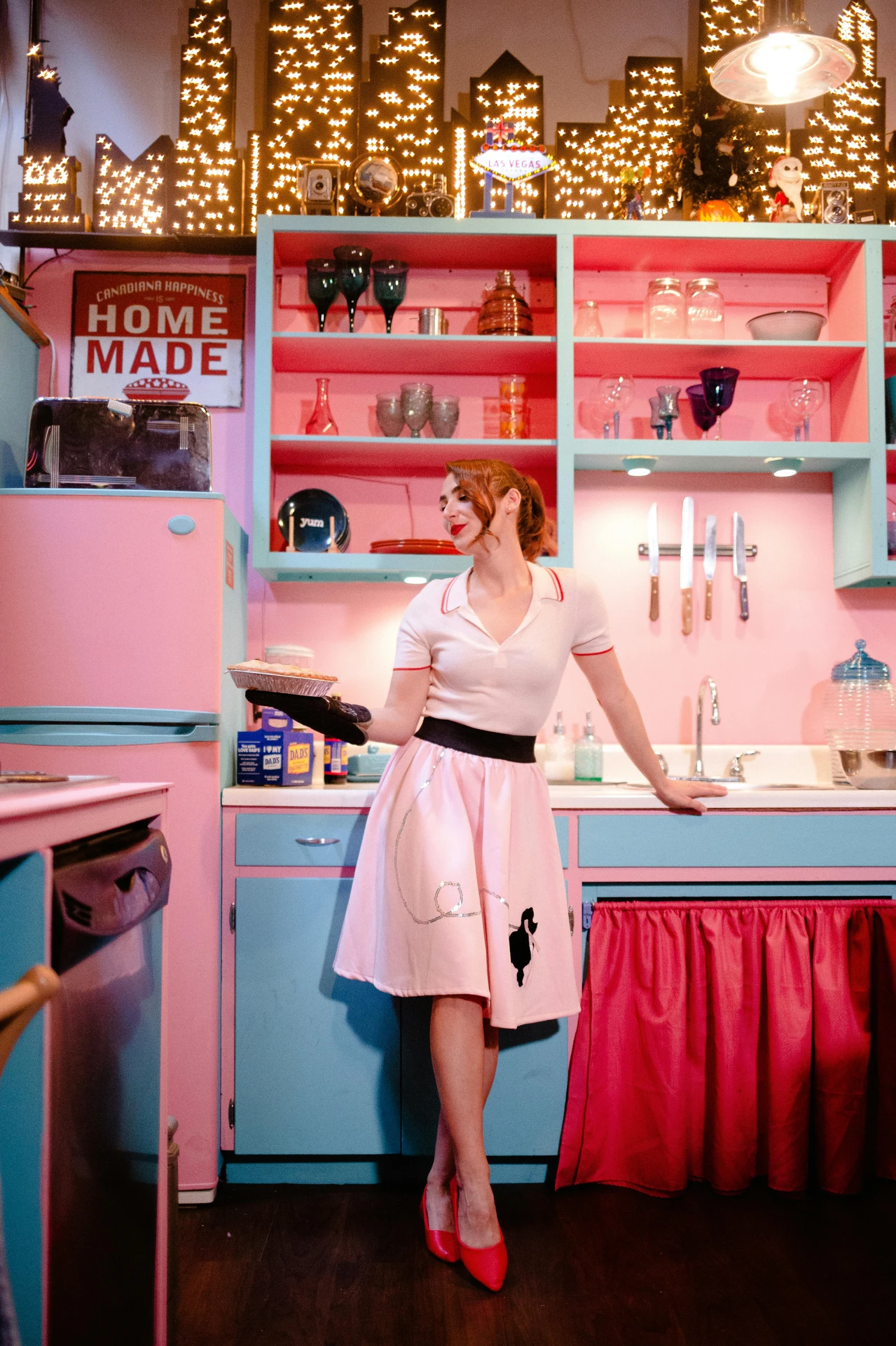 Vibrant kitchen styled with a bold retro look, featuring pastel pink and turquoise cabinets, vintage appliances, and playful open shelving with colorful glassware. A woman in a classic poodle skirt holds a freshly baked pie, adding to the nostalgic charm. While there's no traditional island, the creative use of color, accessories, and kitschy details offers unique inspiration for kitchen island decor ideas that embrace retro flair.