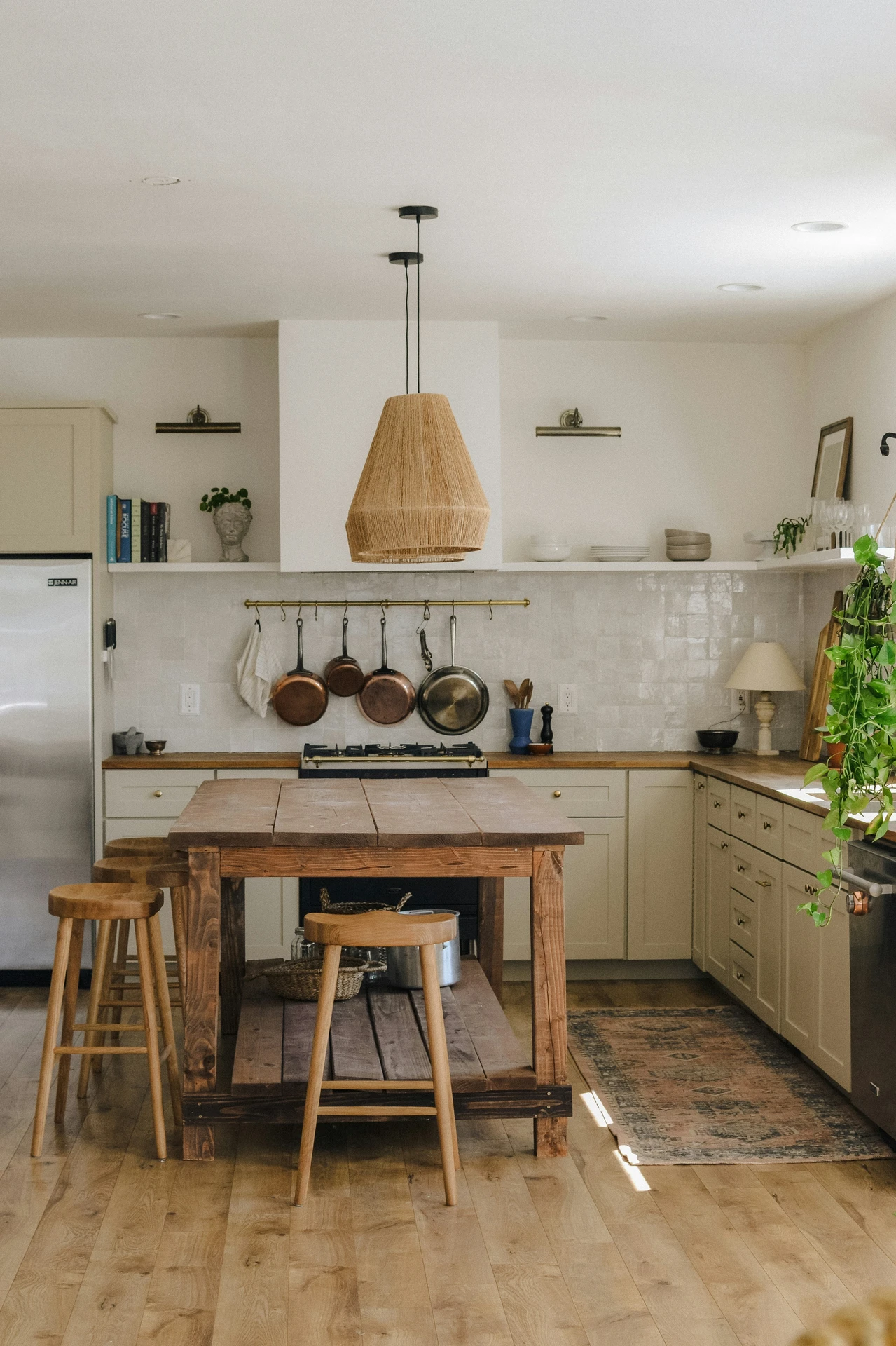 Warm and inviting kitchen featuring a handcrafted wooden rustic kitchen island with natural tones and visible wood grain. The island is paired with simple wooden stools and subtle storage underneath, offering practical and charming kitchen island decor ideas. A woven pendant light, hanging copper pans, and open shelving add to the cozy, farmhouse-inspired aesthetic.