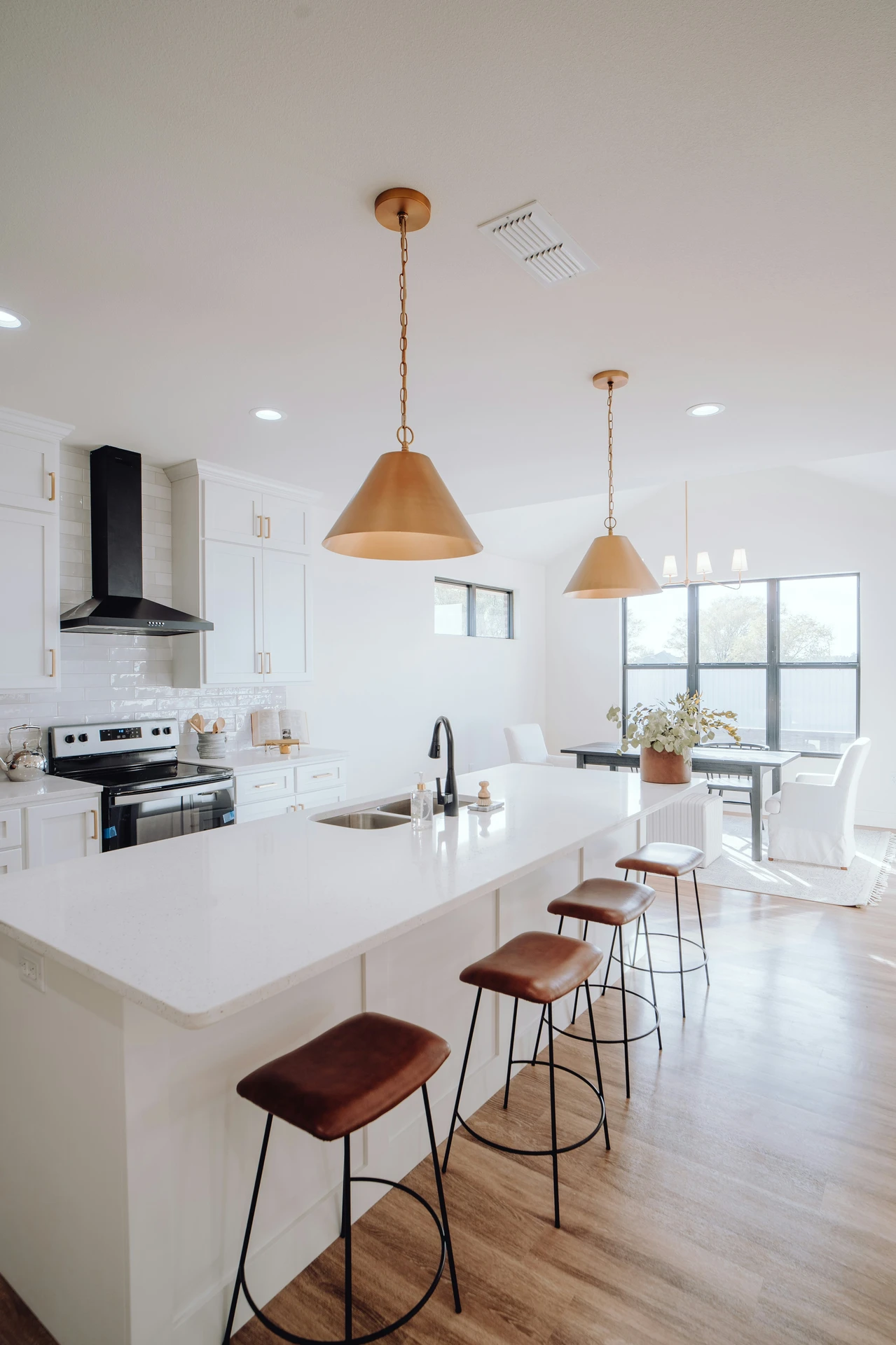 Bright and airy kitchen featuring a sleek white island with modern black fixtures and minimalistic kitchen island decor ideas, including a vase with neutral-toned greenery. Four leather kitchen counter stools with black metal frames provide stylish seating, while oversized gold pendant lights add warmth and elegance to the contemporary space.
