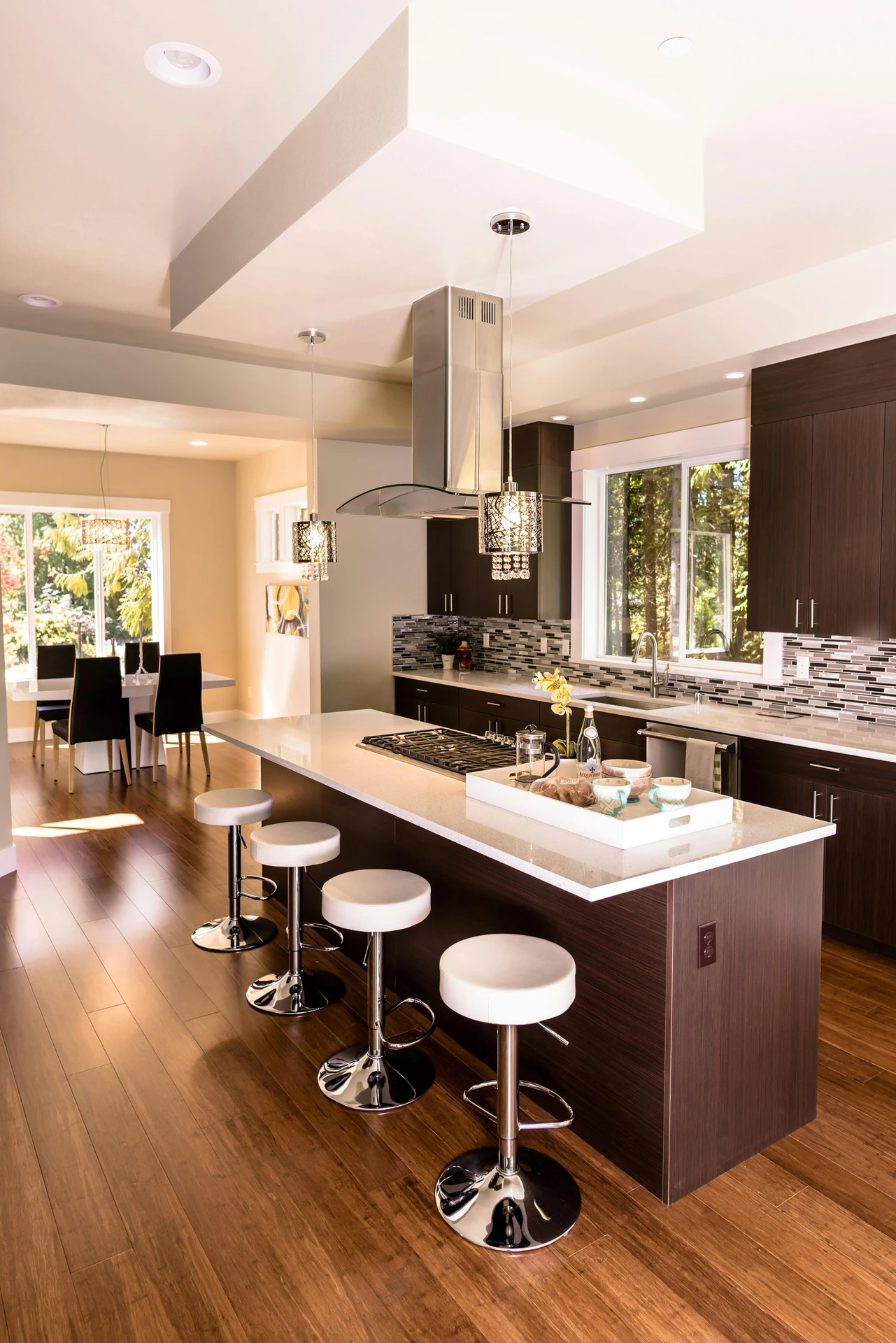 Elegant open-concept kitchen featuring a spacious island with sleek dark wood cabinetry and white countertops. The island is adorned with a decorative tray holding bowls, a flower vase, and a bottle, offering refined kitchen island decor ideas. Modern pendant lighting and chrome bar stools complete the contemporary look, blending style and functionality.