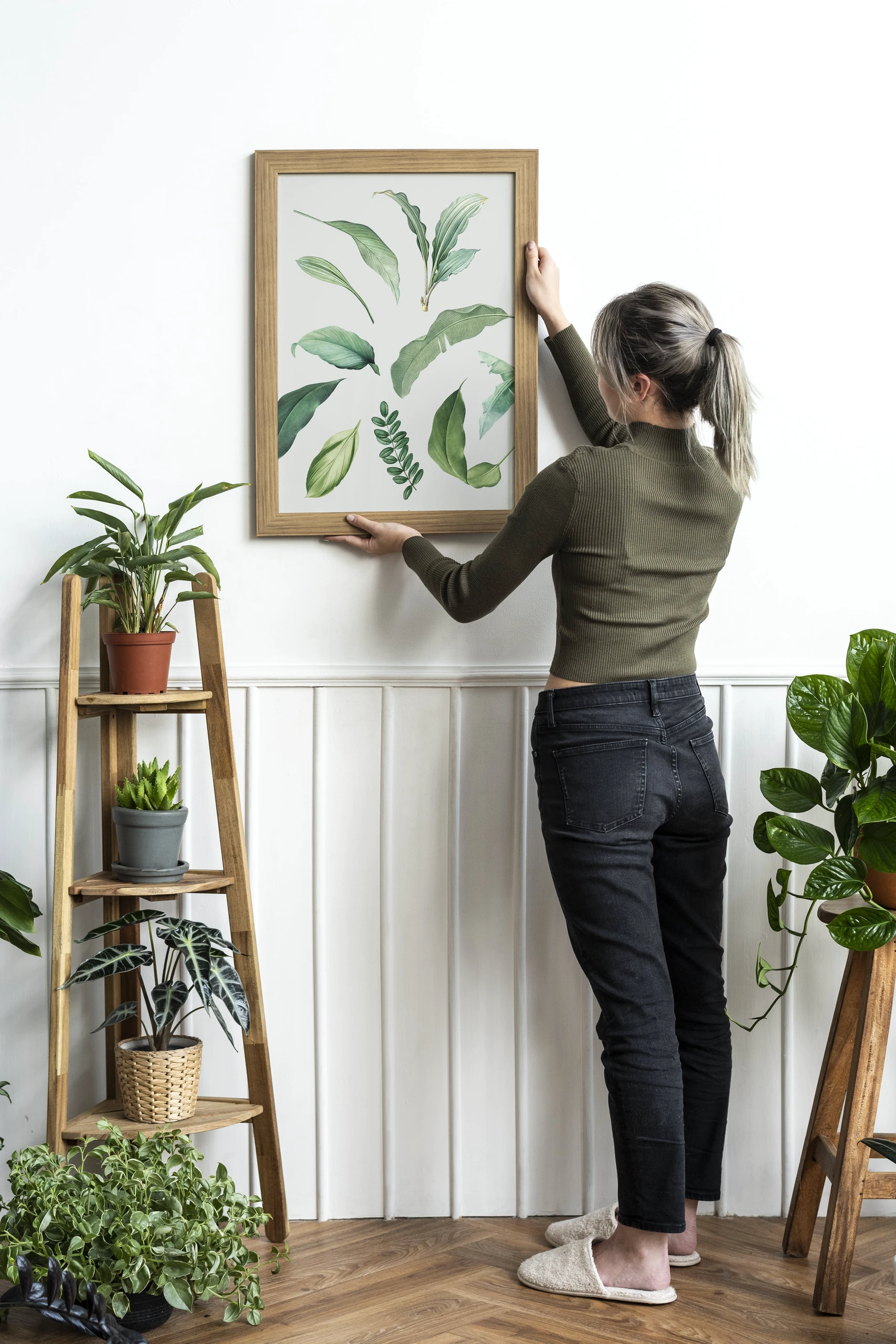 Woman decorating a white living room wall with a framed nature print featuring green leaves, surrounded by indoor plants. An inspiring example of living room decor ideas for walls using botanical and nature prints.