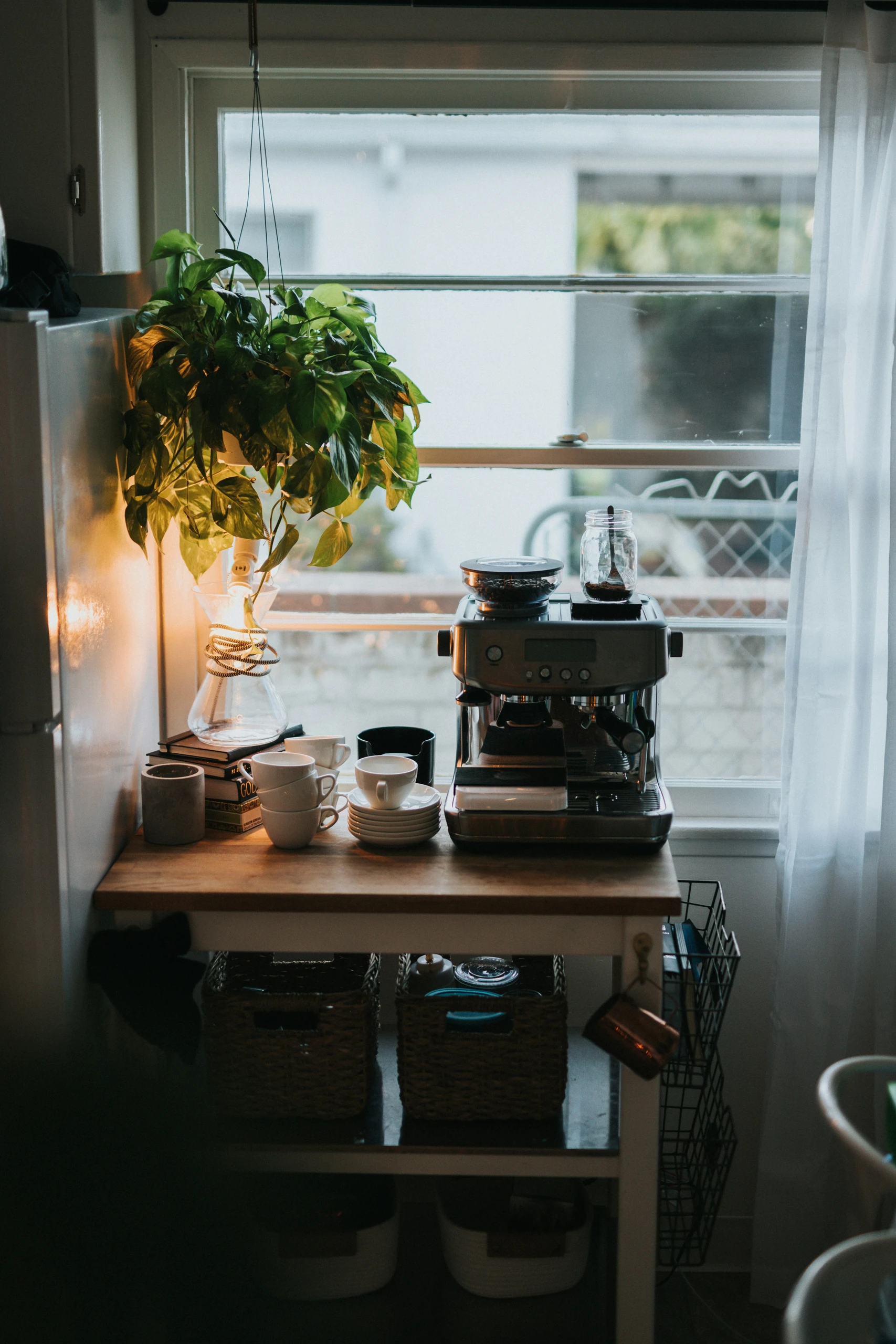 Cozy coffee station setup by the window featuring a sleek espresso machine, stacked mugs, and hanging greenery. An inviting example of small kitchen decor ideas that make room for a stylish and functional coffee station.