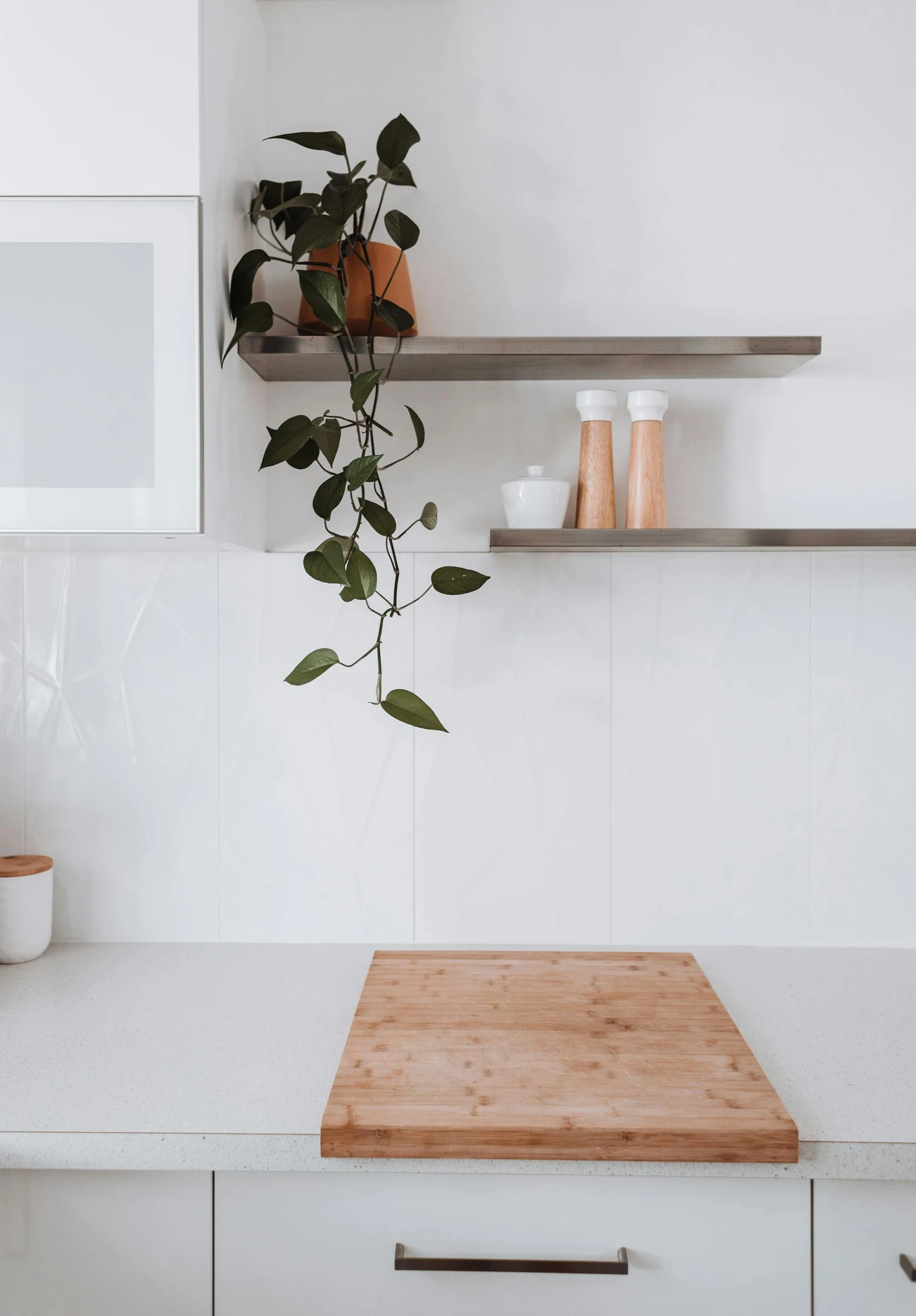 Minimalist white kitchen showcasing small kitchen decor ideas with sleek floating shelves above the counter, holding wooden salt and pepper mills, a white jar, and a potted trailing plant for a touch of greenery.