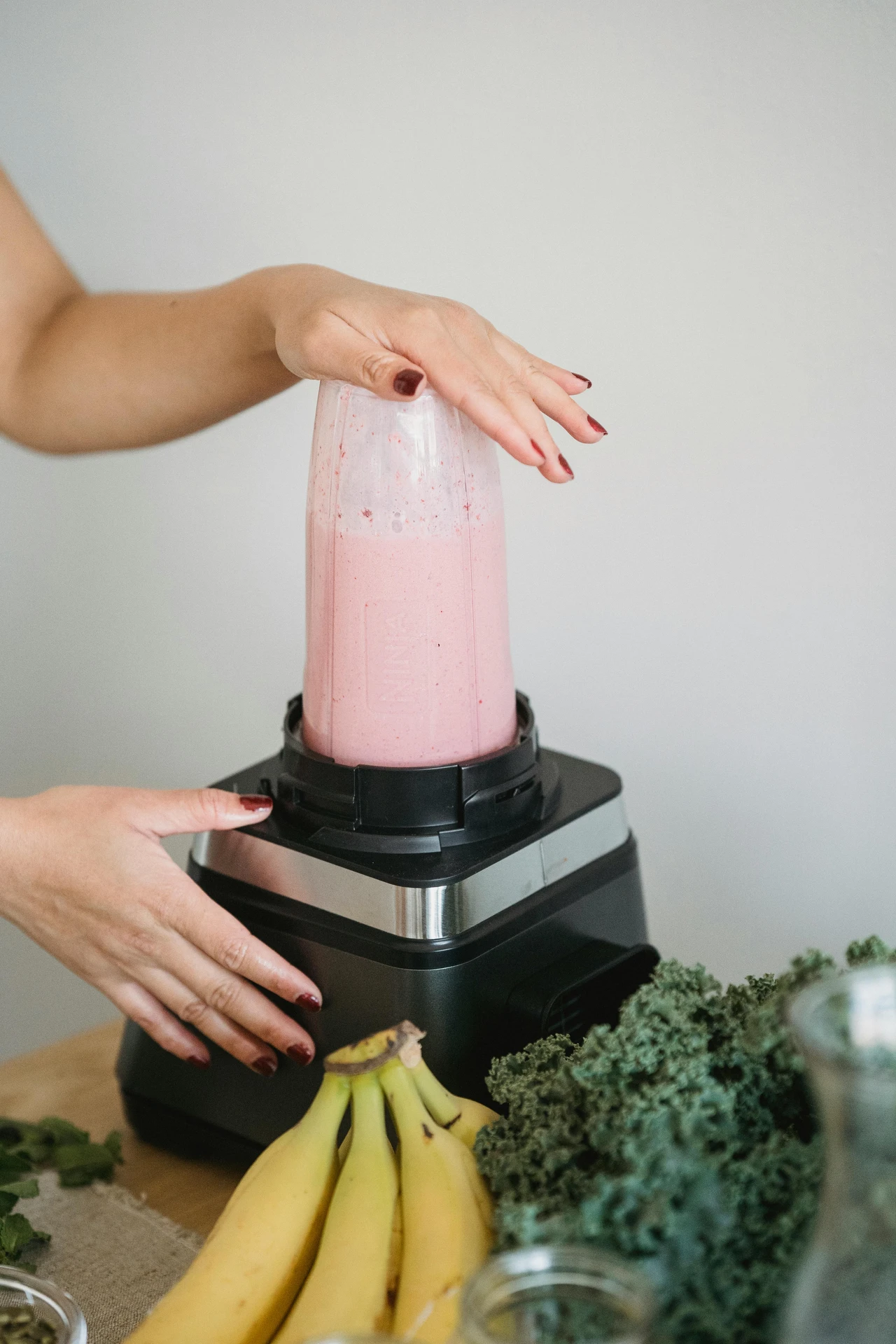 A person using a compact blender, one of the best small kitchen appliances, to prepare a pink smoothie. The scene features fresh ingredients including bananas, kale, and herbs on a wooden surface, highlighting a versatile setup for making healthy drinks and recipes in any home kitchen.