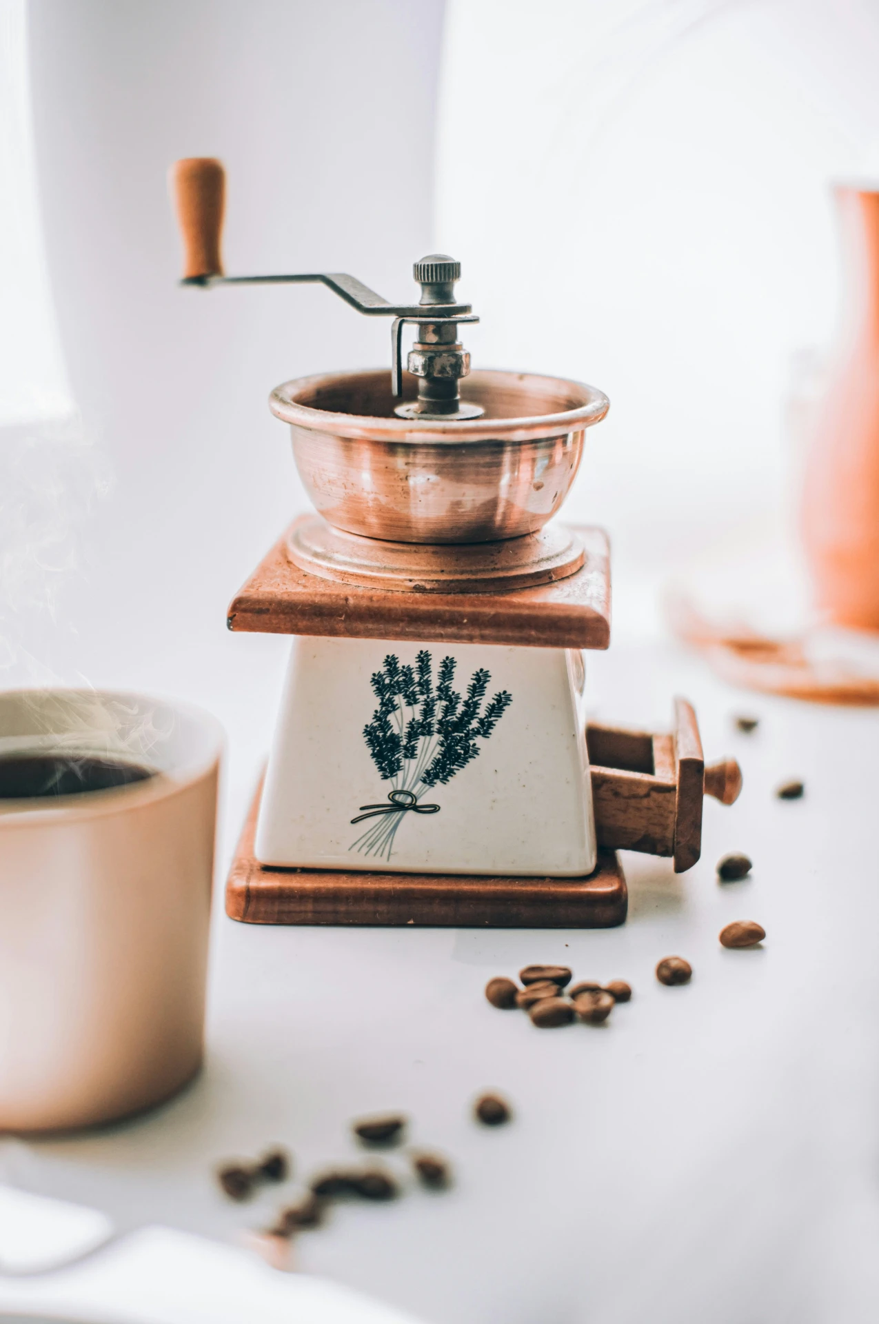 Charming vintage-style coffee grinder, a timeless example of small kitchen appliances that blends functionality with rustic decor. Featuring a ceramic base with a lavender motif, a copper grinding bowl, and a wooden handle, this coffee grinder is surrounded by scattered coffee beans and a steaming cup, perfect for crafting fresh grounds for a variety of brewing methods.