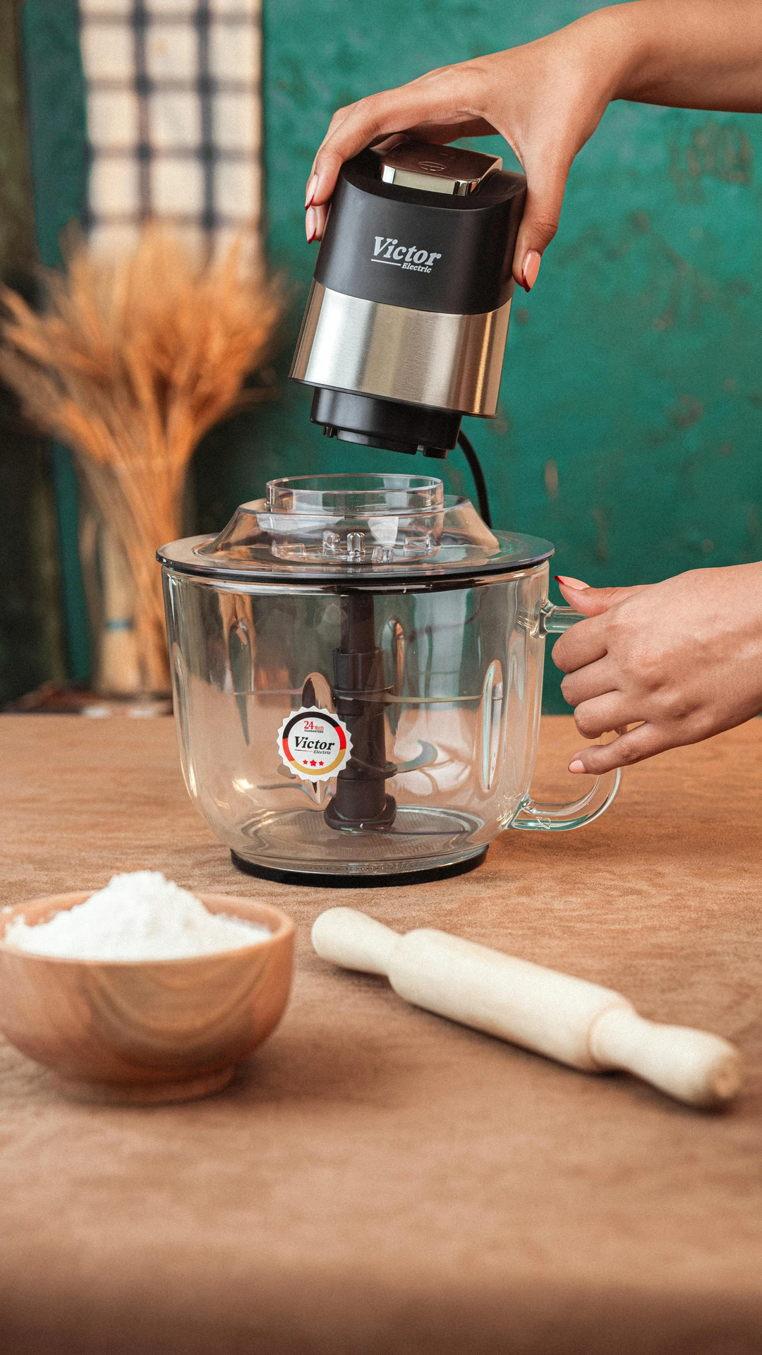 A sleek food processor featuring a clear glass mixing bowl and a stainless steel motor unit, showcased as part of small kitchen appliances. The appliance is held by a person preparing it for use, surrounded by baking essentials like a rolling pin and a bowl of flour, making it ideal for chopping, mixing, or kneading in both everyday cooking and creative culinary projects.
