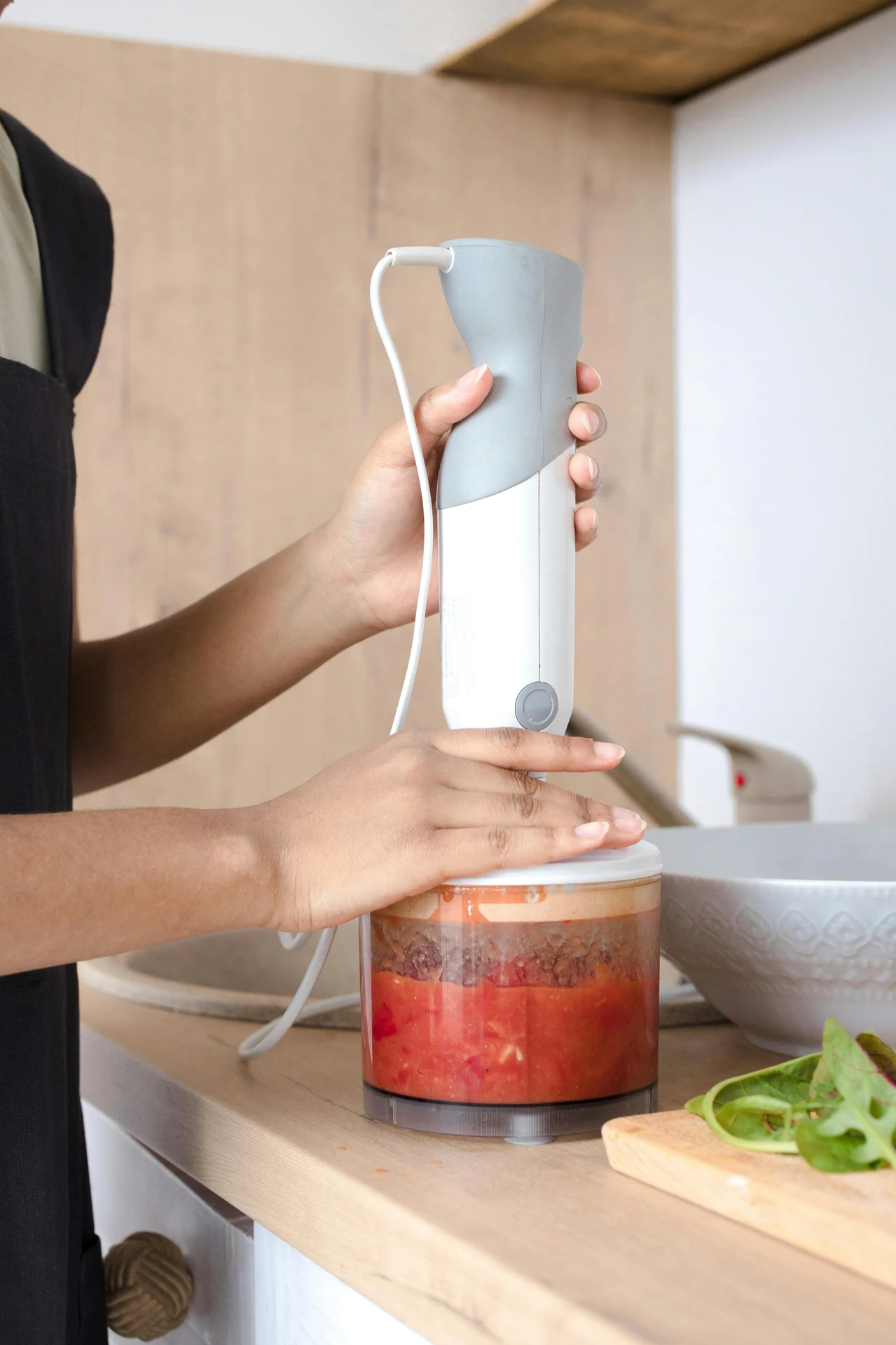 Person using a hand blender, a versatile member of small kitchen appliances, to puree a mixture of vegetables and other ingredients in a transparent container. The appliance's ergonomic grip and compact size make it ideal for preparing soups, sauces, smoothies, and more, while fresh greens on a cutting board nearby hint at healthy and creative cooking possibilities.