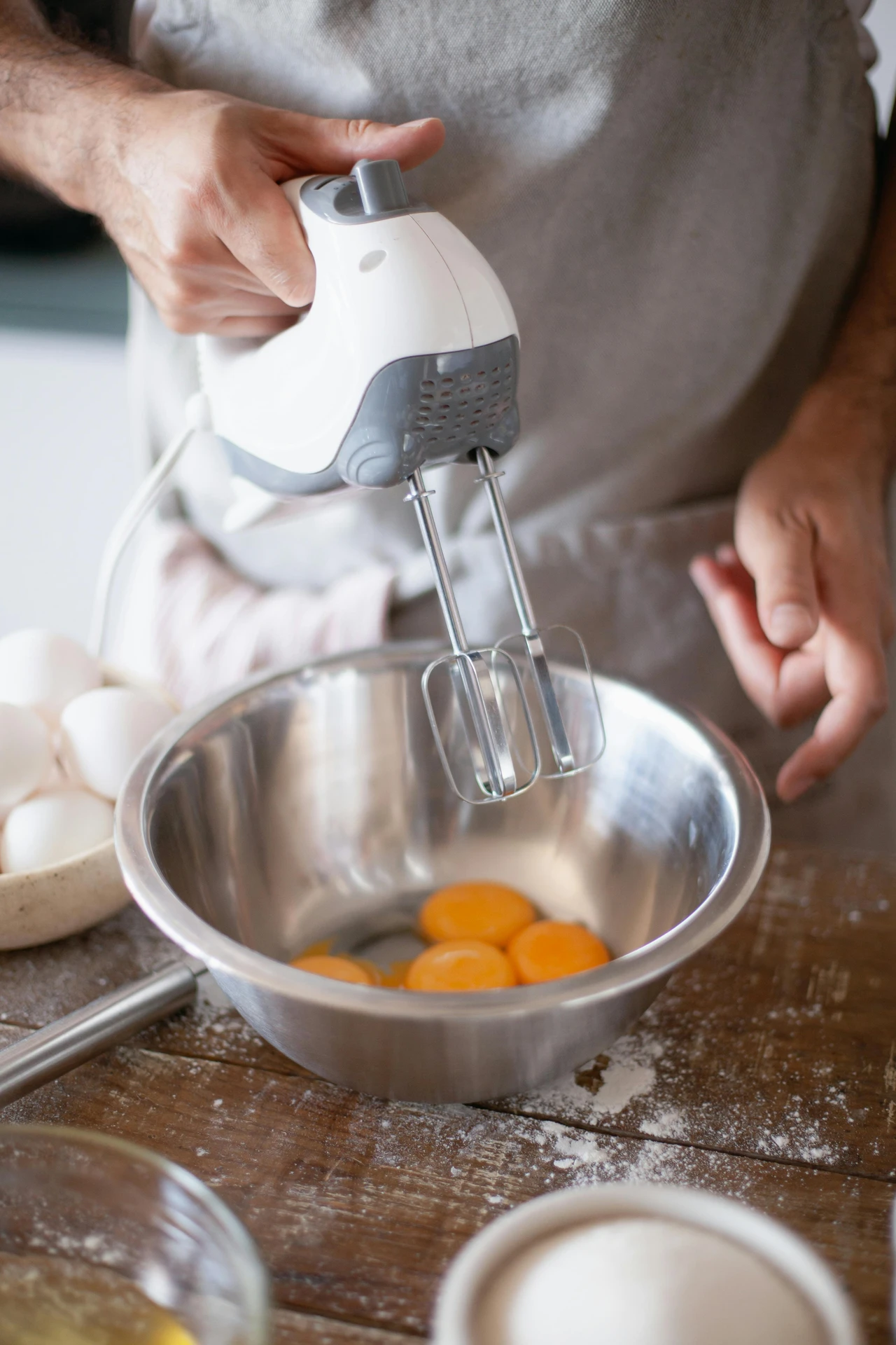 A person holds a hand mixer above a stainless steel mixing bowl containing several egg yolks, ready to blend ingredients for baking or cooking. Surrounded by eggs, flour, and other kitchen essentials, this scene highlights how small kitchen appliances like a hand mixer make it easy to prepare everything from cakes and pastries to sauces and whipped toppings.