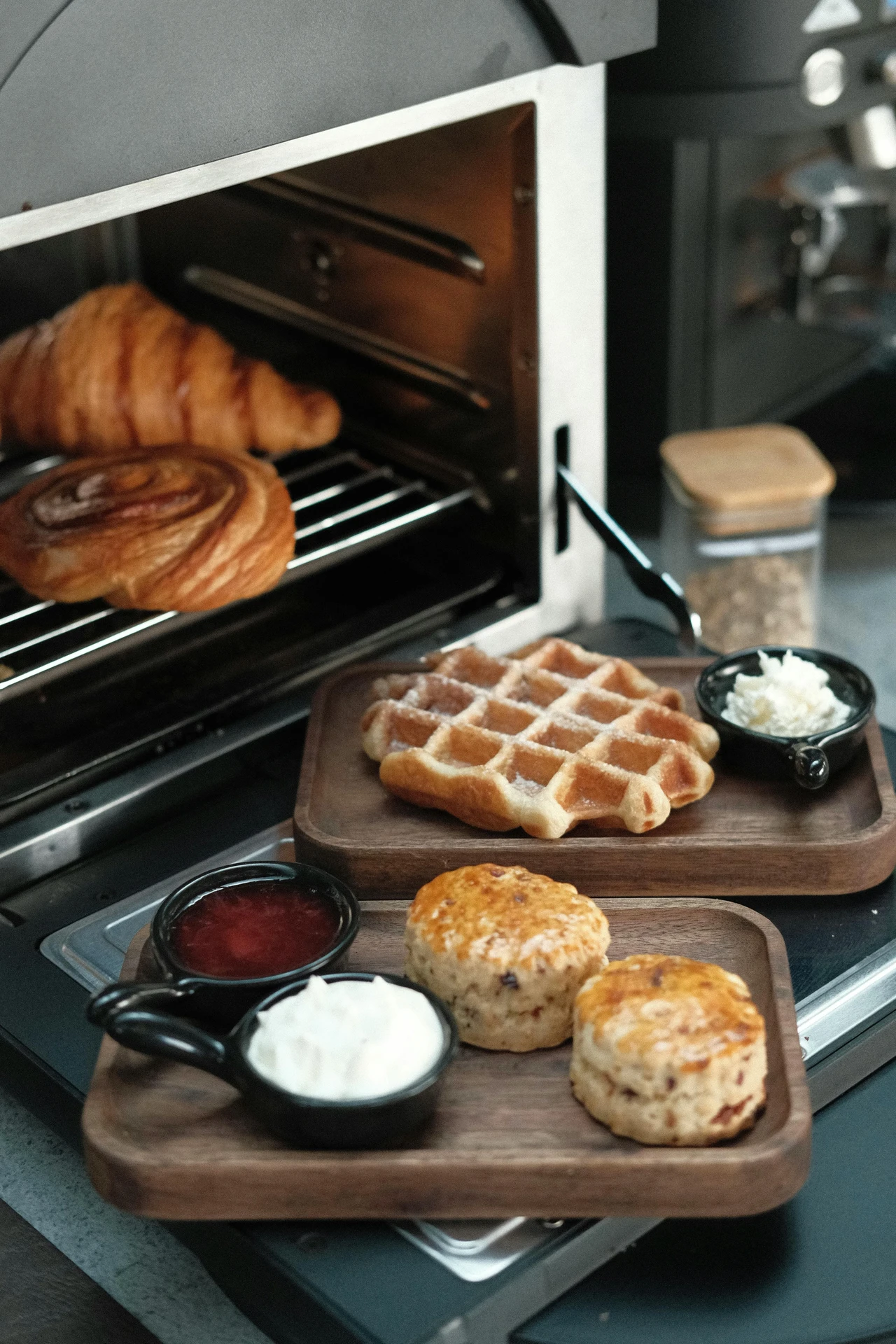 A compact toaster oven, one of the most versatile small kitchen appliances, warming golden croissants and a cinnamon roll, with a wooden serving tray nearby holding a sugar-dusted waffle, fresh scones, whipped cream, and fruit jam, perfect for preparing anything from breakfast pastries to savory snacks.