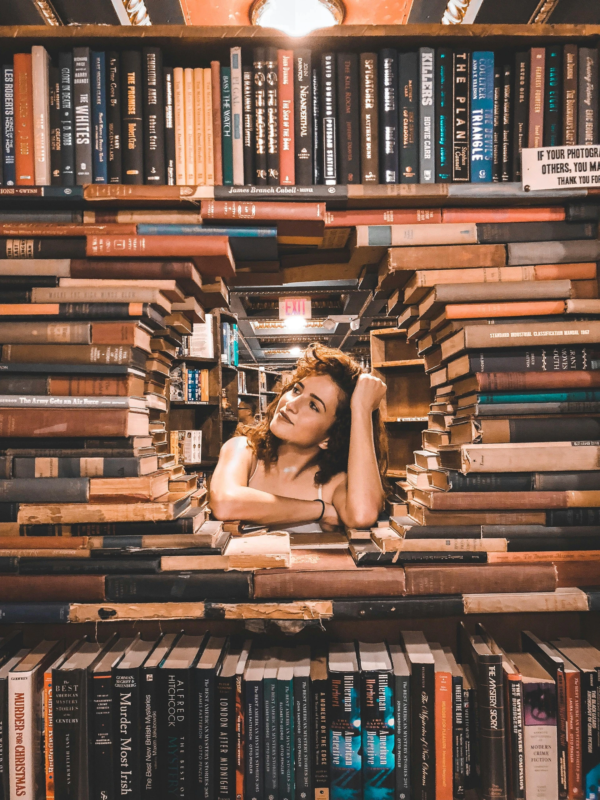 Creative bookshelf decorating ideas brought to life with building shapes of books. A young woman poses playfully within a tunnel-like archway sculpted entirely out of stacked books, blending art and literature in a captivating way. Surrounded by colorful spines and well-worn pages, the scene combines vintage charm with imaginative design, showing how books can be used not only for reading but as striking architectural decor in any book lover's space.