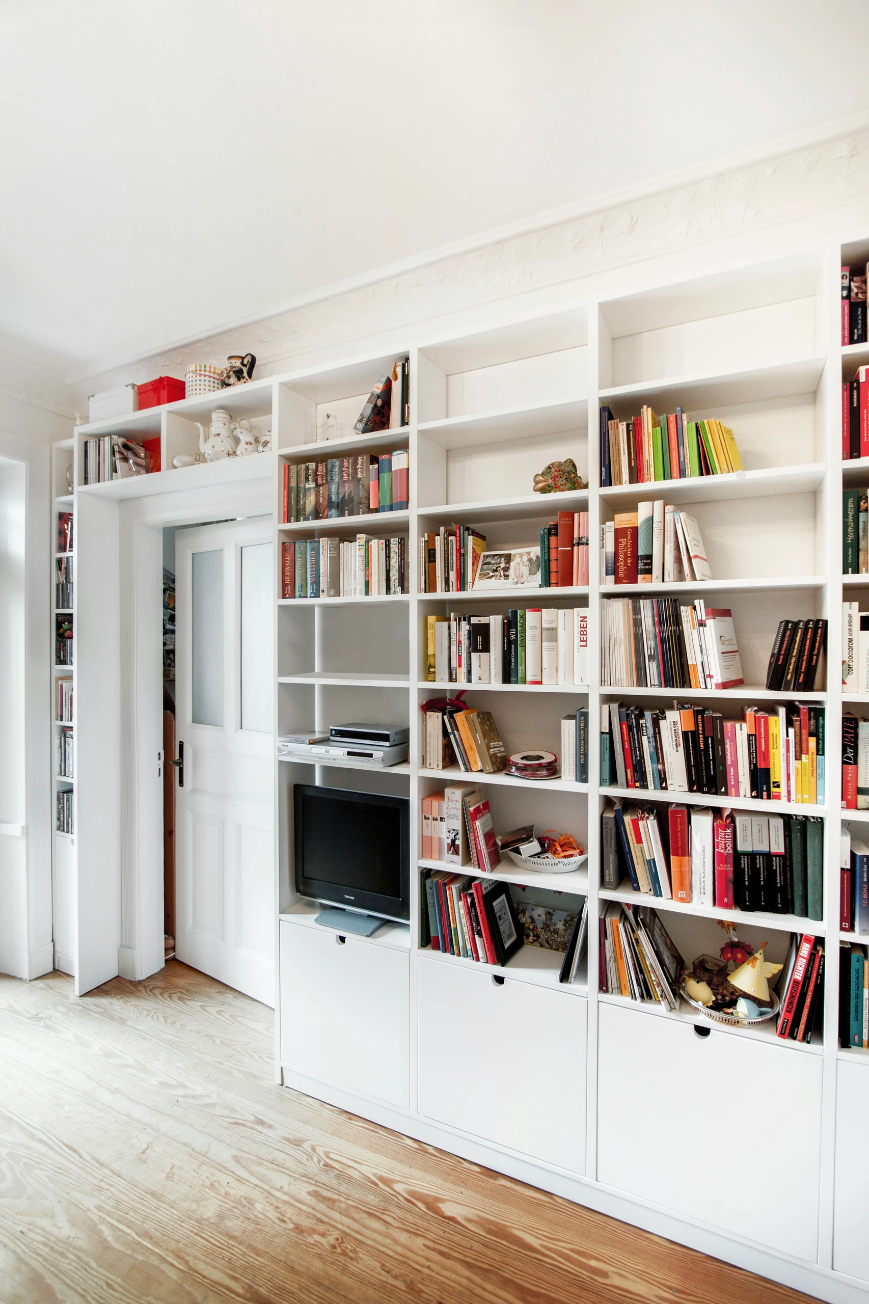 An inspiring example of bookshelf decorating ideas focused on maximized storage. This built-in white shelving unit efficiently frames a doorway, stretching from floor to ceiling and wall to wall, and combines open and closed compartments for books, décor, media devices, and personal treasures. The neatly arranged books, ceramics, and colorful accents create a vibrant yet functional look, perfect for small spaces, home offices, or multifunctional living areas seeking both style and practicality.
