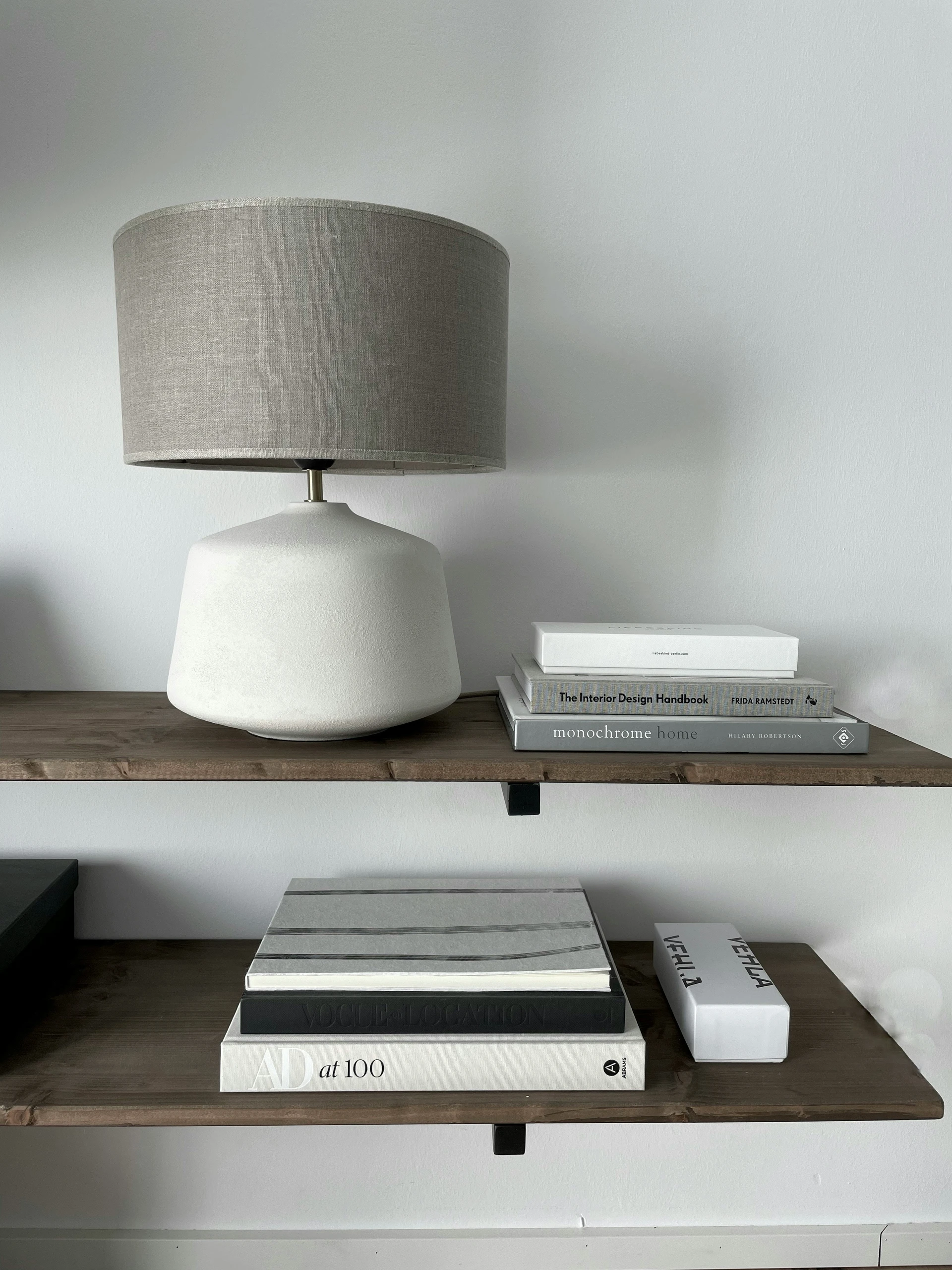 Minimalist bookshelf decorating ideas in a monochrome style, featuring two wooden floating shelves adorned with neutral-toned coffee table books, a sculptural white table lamp, and a sleek rectangular box labeled 