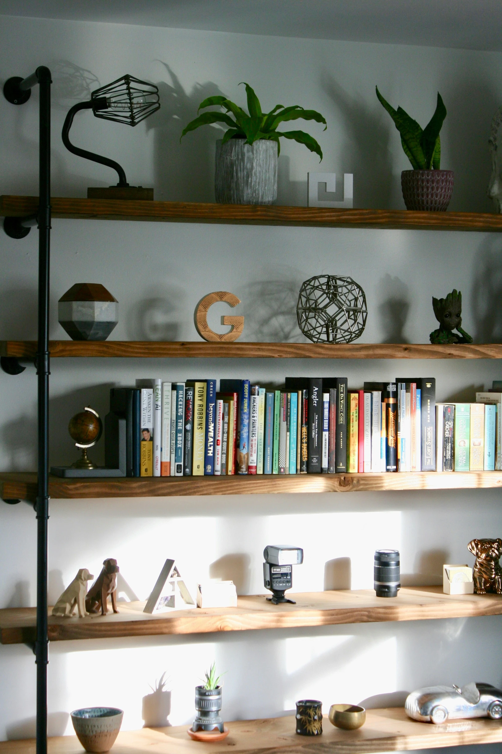 A creative example of bookshelf decorating ideas featuring a personalized bookshelf styled with a mix of greenery, books, travel mementos, and unique decor pieces. The shelves display potted plants, wooden letters, geometric sculptures, and pop culture figurines alongside a curated book collection. Additional shelves below showcase personal touches like photo frames, camera equipment, ceramic bowls, and miniature sculptures, offering a versatile and lived-in feel that reflects the owner's personality and interests.