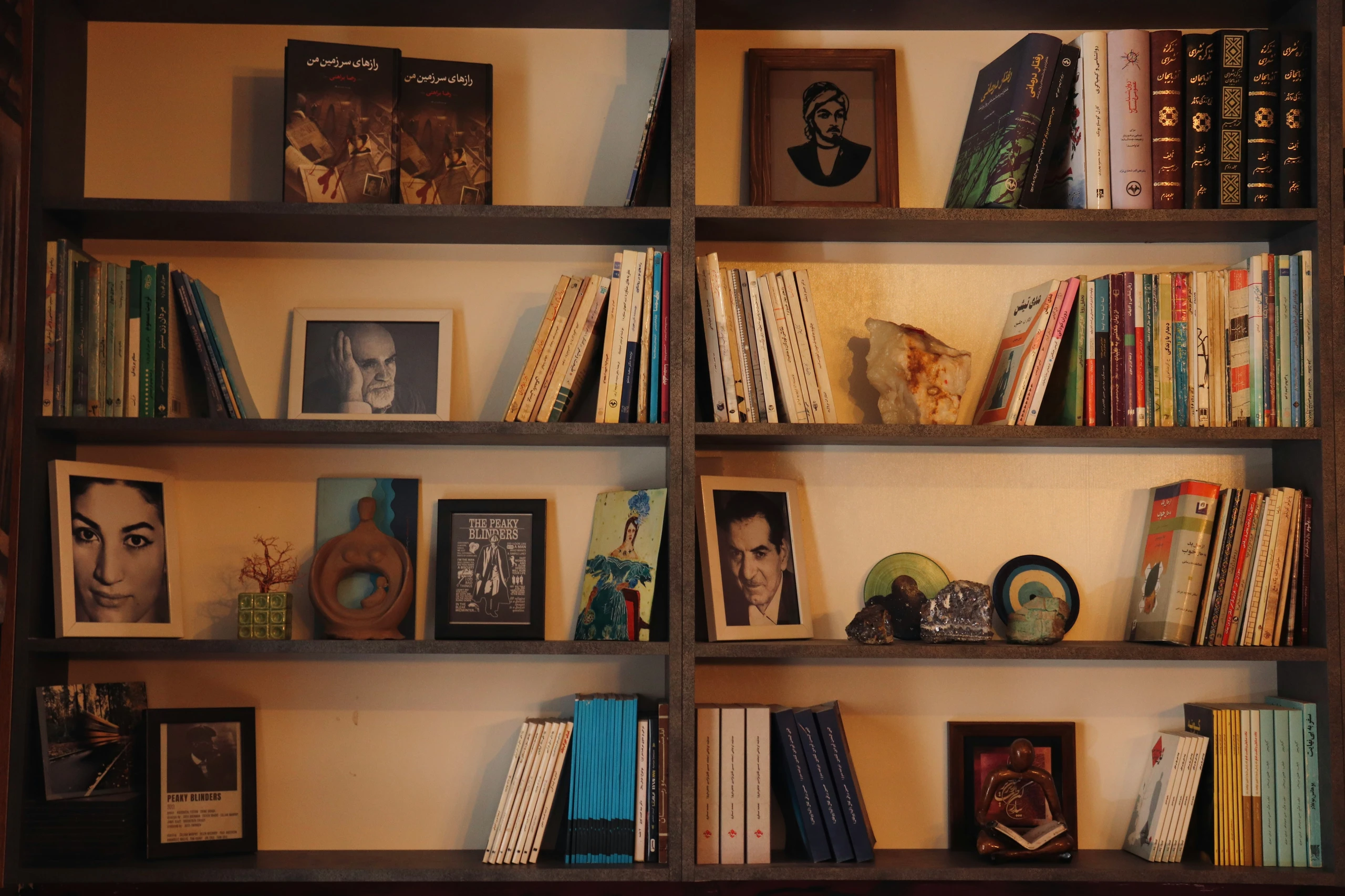 A cozy and personalized bookshelf filled with creative bookshelf decorating ideas, blending literature with meaningful decor. The shelves feature an eclectic mix of books in various colors and sizes, accented by framed black-and-white photo portraits and artistic prints. Natural elements like a large quartz crystal and unique figurines add texture, while thoughtful touches such as mini sculptures and curated photo frames create a warm, expressive ambiance. Ideal for anyone seeking versatile styling inspiration.