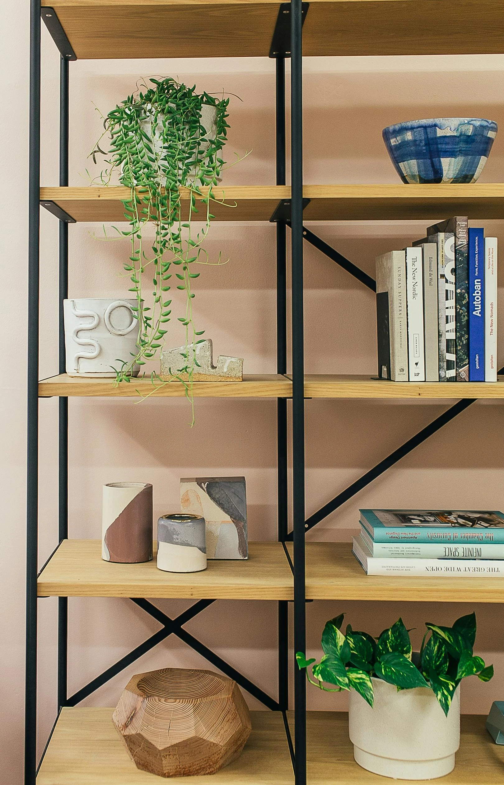 A modern open bookshelf styled with creative bookshelf decorating ideas, featuring a balanced mix of plants, candles, and bowls. Cascading greenery and a potted plant add a fresh touch, while sculptural ceramics and minimalist candles bring texture and personality. Art books and stacked volumes provide a grounded feel, and a wooden geometric bowl anchors the arrangement with natural warmth, a perfect inspiration for a stylish and versatile shelving setup.