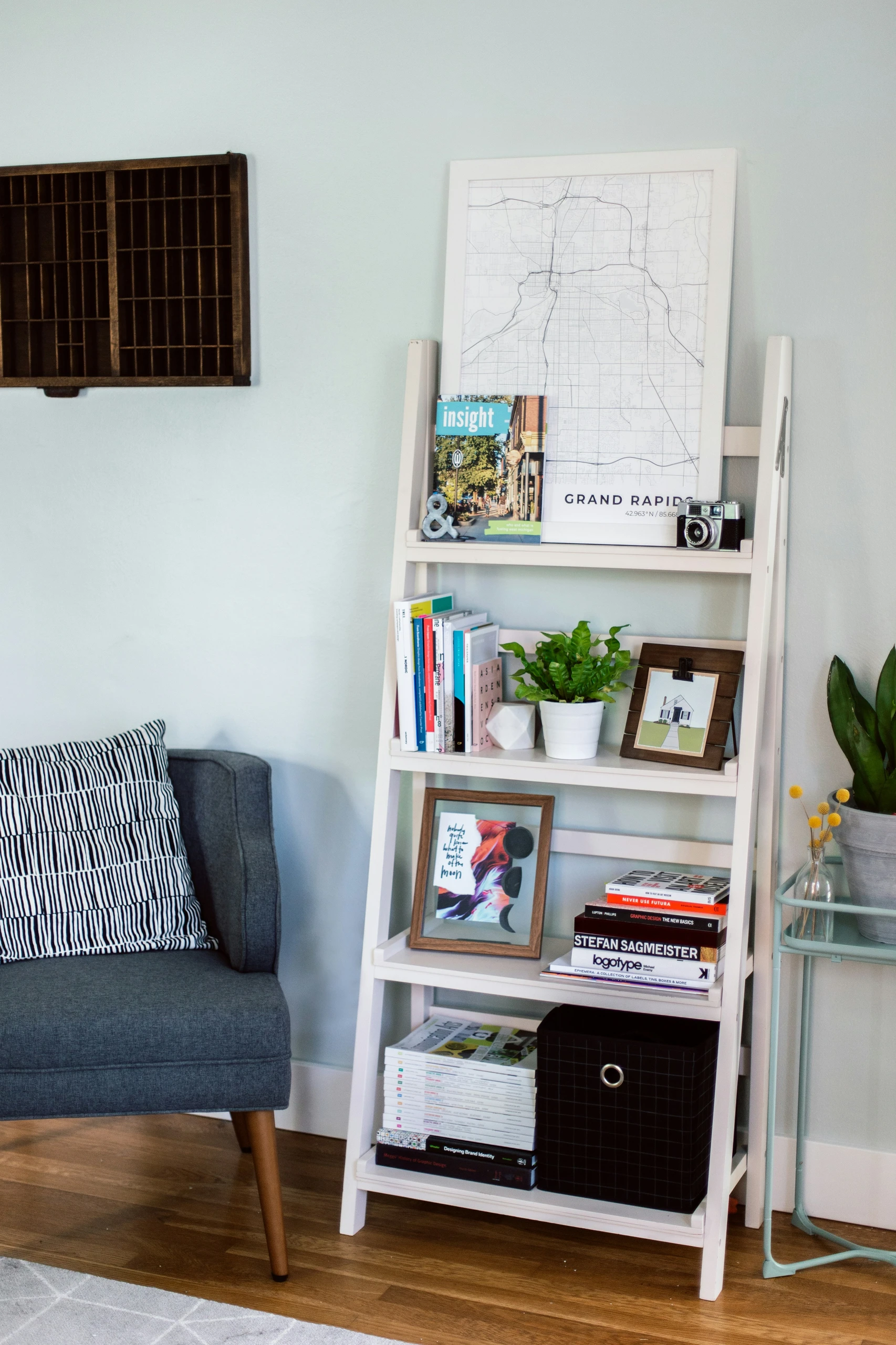 A stylish example of bookshelf decorating ideas featuring a small ladder-shaped bookshelf in a modern living space. The white, tiered shelving unit leans against a pale blue wall and showcases a creative mix of books, framed art, a potted plant, a vintage camera, and a black storage box. Each shelf combines functional storage with personal décor elements, offering a compact yet visually engaging solution perfect for small apartments, reading nooks, or minimalist interiors.