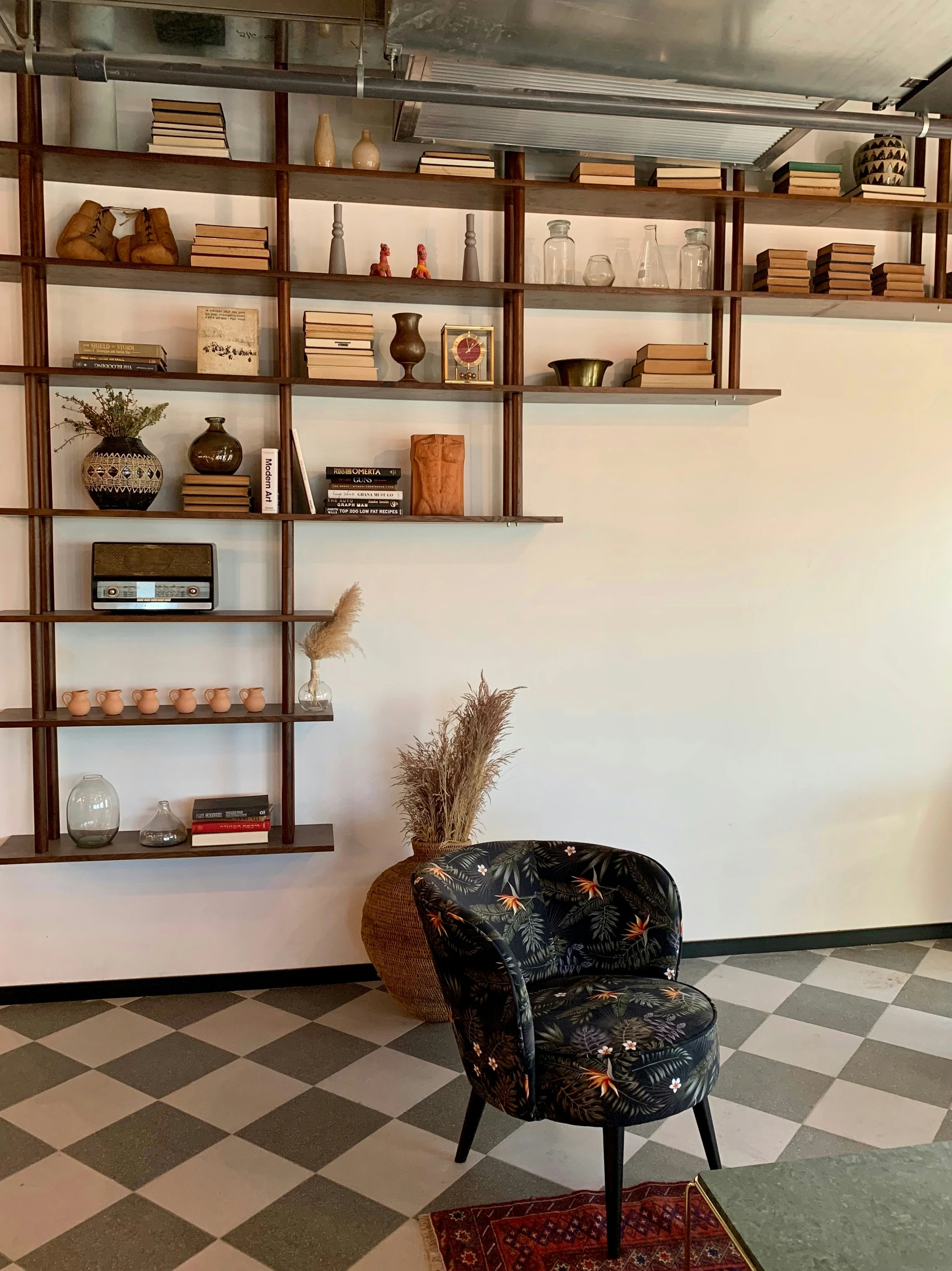A stylish example of bookshelf decorating ideas featuring a special shape wall-mounted shelving unit that combines form and function. The open, asymmetrical structure displays a mix of stacked books, ceramics, vintage decor, and glassware, creating a curated and artistic look. Neutral tones and warm wood contrast with a patterned accent chair and checkered floor, making the space feel both versatile and cozy. Ideal for modern or boho-inspired interiors.