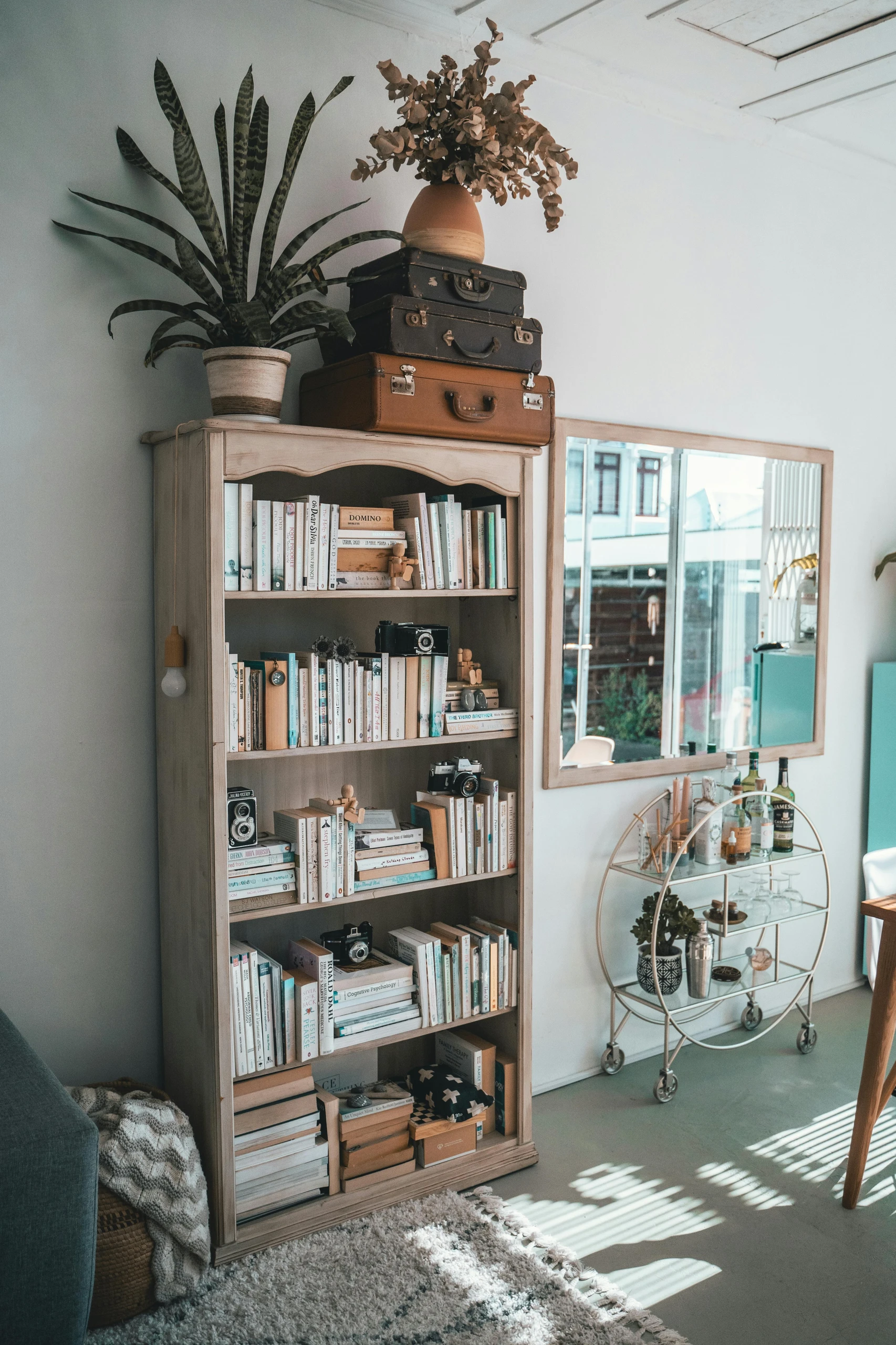 A cozy and stylish corner featuring bookshelf decorating ideas, with a tall wooden bookshelf filled with neutral-toned books, vintage cameras, and small wooden art figures. On top, stacked vintage suitcases and a terracotta vase with dried foliage add character and height. A large leafy plant adds a fresh touch, while a bar cart with glassware and spirits sits nearby, reflecting a well-curated yet lived-in aesthetic.