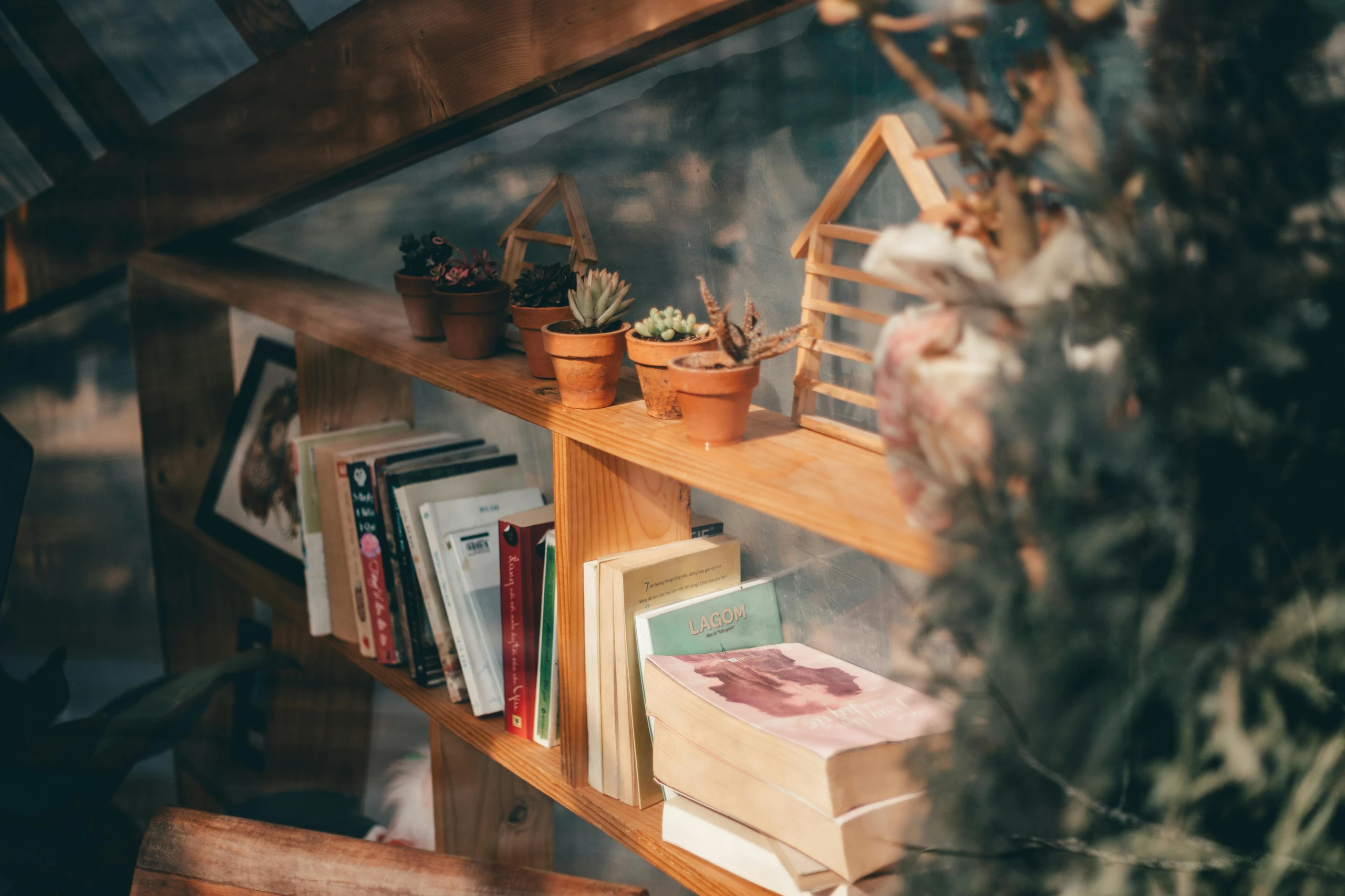 A cozy wooden bookshelf styled with versatile bookshelf decorating ideas, featuring a row of small terracotta pots filled with assorted succulents and cacti on the upper shelf. Below, a curated mix of books, some stacked and some upright, adds a casual and inviting vibe. Decorative wooden house-shaped accents and framed art tucked behind the books bring warmth and personality to this charming corner.