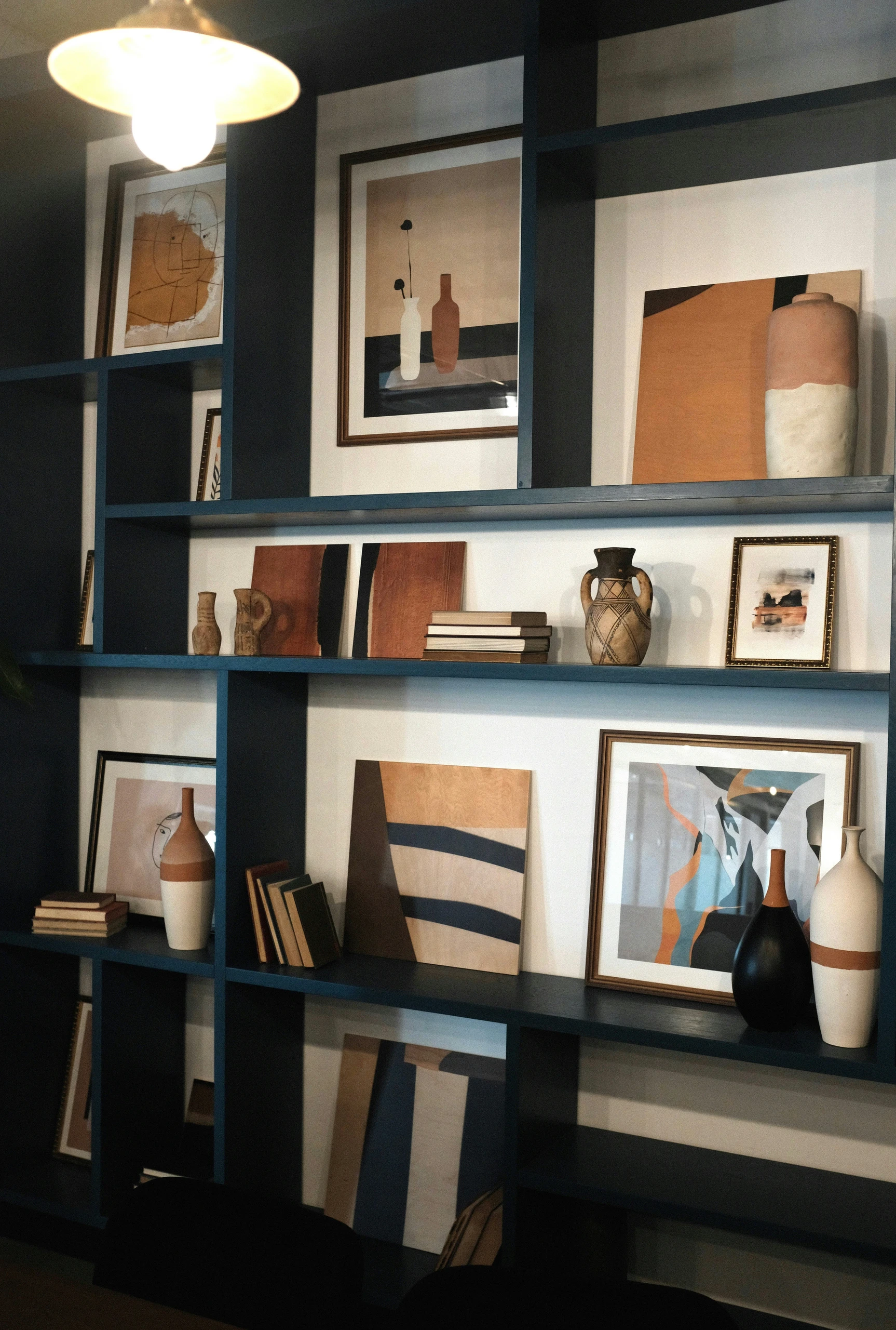 Elegant bookshelf decorating ideas using uniform shapes and colors, featuring a navy-blue shelving unit adorned with neutral-toned abstract art, stacked books in earthy hues, and ceramic vases in complementary shades of cream, terracotta, and black. The cohesive color palette and repeated forms create a calm, curated look that suits modern, boho, or minimalist interiors alike.