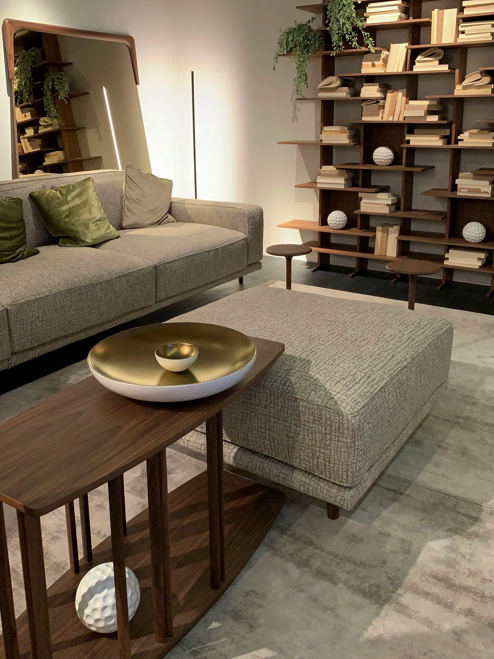 A modern living room showcasing creative bookshelf decorating ideas with a striking wooden shelving unit featuring both vertically and horizontally arranged books. The geometric layout adds visual rhythm, while cascading green plants and sculptural white décor spheres provide organic contrast. Neutral-toned furniture, a textured ottoman, and a sleek accent mirror contribute to a calm, elegant ambiance, making this setup ideal for anyone looking to combine functional storage with bold, artful interior design.