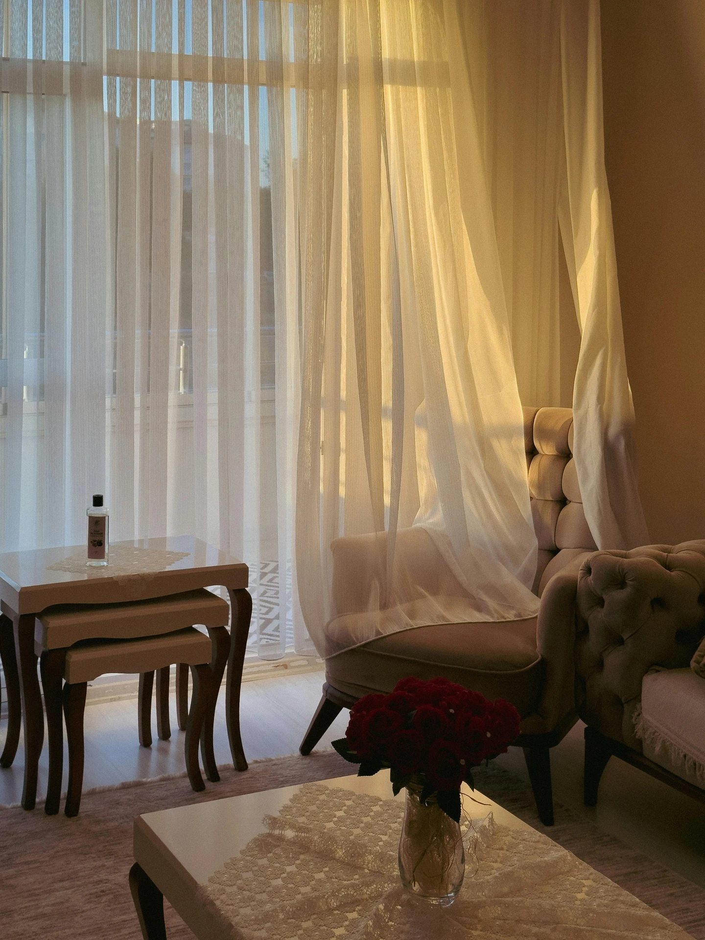 Elegant curtain ideas for the living room with sheer drapes in a delicate white touch, softly flowing in the sunlight. The curtains frame a cozy space with a tufted armchair, nesting tables, and a coffee table adorned with lace and a vase of red roses, creating a warm and versatile balance of light and comfort.