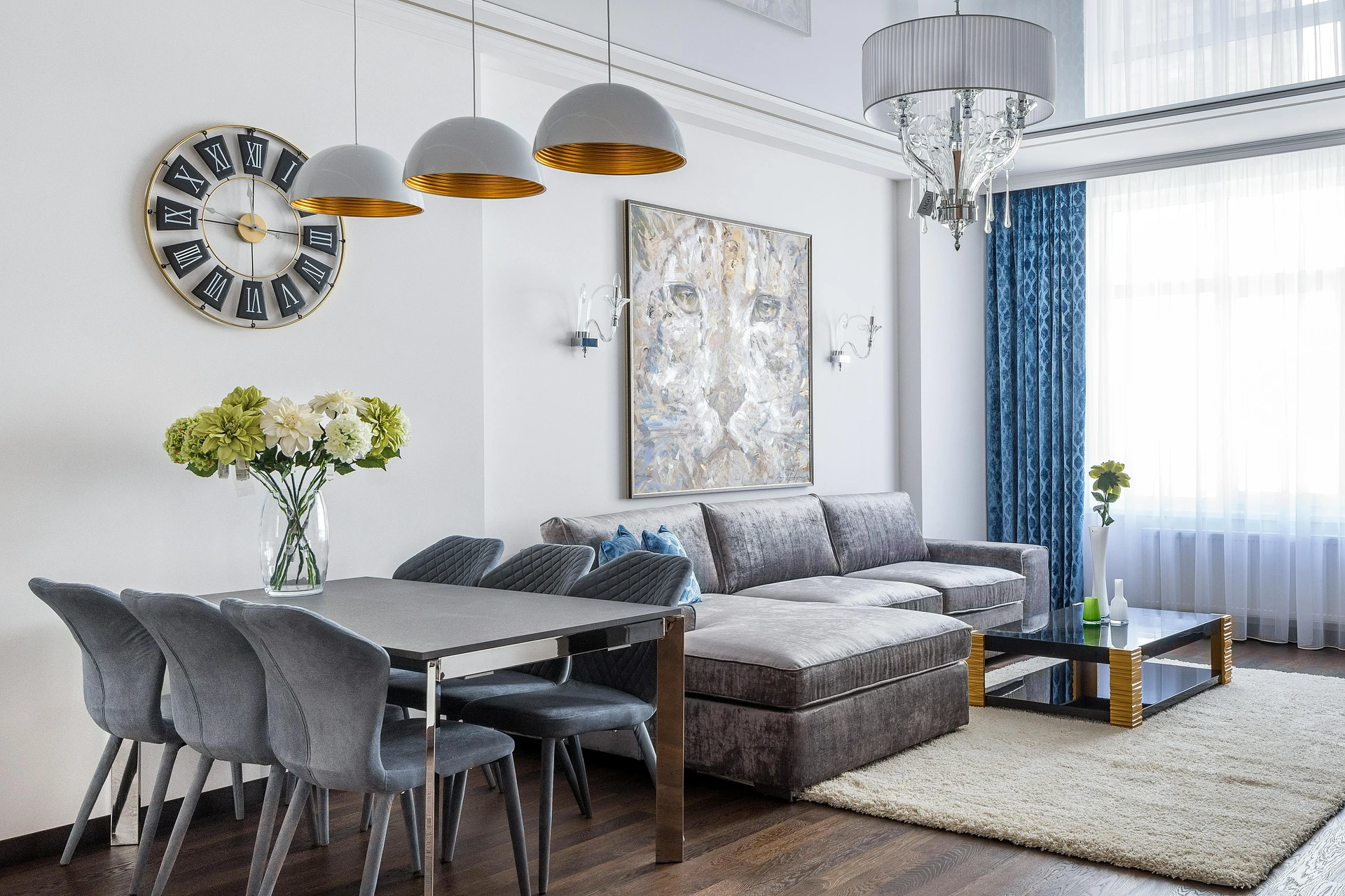 Modern curtain ideas for the living room featuring elegant drapes in a rich blue pattern paired with sheer white curtains, bringing depth and sophistication to the space. The room includes a plush gray sectional sofa, a sleek black-and-gold coffee table, and a dining set with velvet chairs, accented by statement lighting and wall art for a versatile and stylish look.