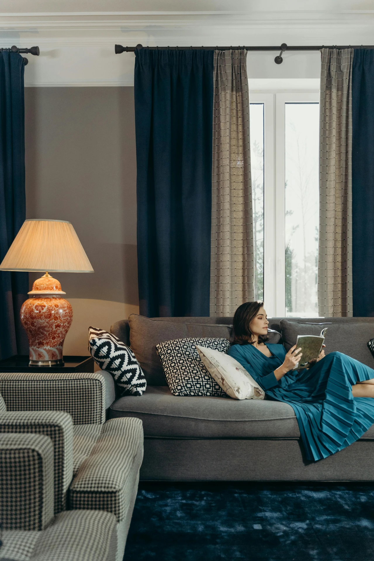 Stylish curtain ideas for the living room featuring two layers: colorful and pattern. Deep blue curtains frame the window while a second layer of patterned beige drapes adds texture and elegance. The cozy space includes a grey sofa with mixed patterned cushions, a woman reading, a vintage ceramic lamp, and a warm, inviting ambiance that feels both modern and timeless.