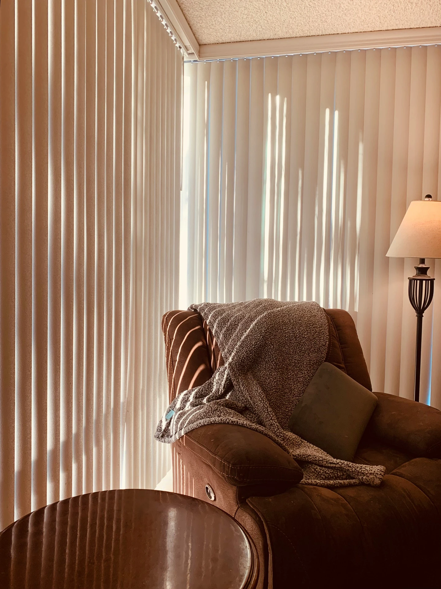Modern curtain ideas for the living room featuring sleek vertical curtain panels in a soft beige tone, allowing light to filter through in elegant stripes. A cozy brown armchair with a textured throw blanket and cushion sits beside a wooden side table and floor lamp, creating a warm and versatile corner perfect for relaxation.