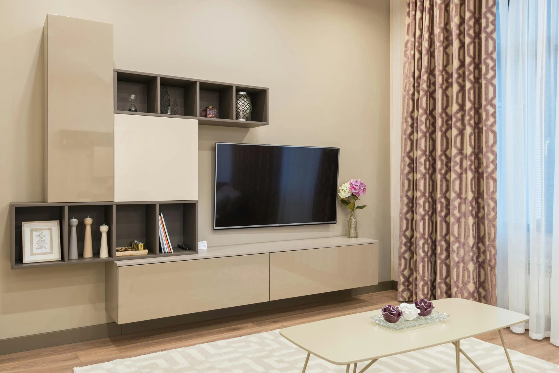Elegant curtain ideas for the living room with patterned drapes in warm beige and muted plum tones, creating a harmonious overall appearance tone in tone with the wall color and furniture. The space features a modern wall-mounted TV unit, decorative shelving with vases and candles, a sleek coffee table, and a neutral rug, all blending seamlessly for a stylish and versatile interior.