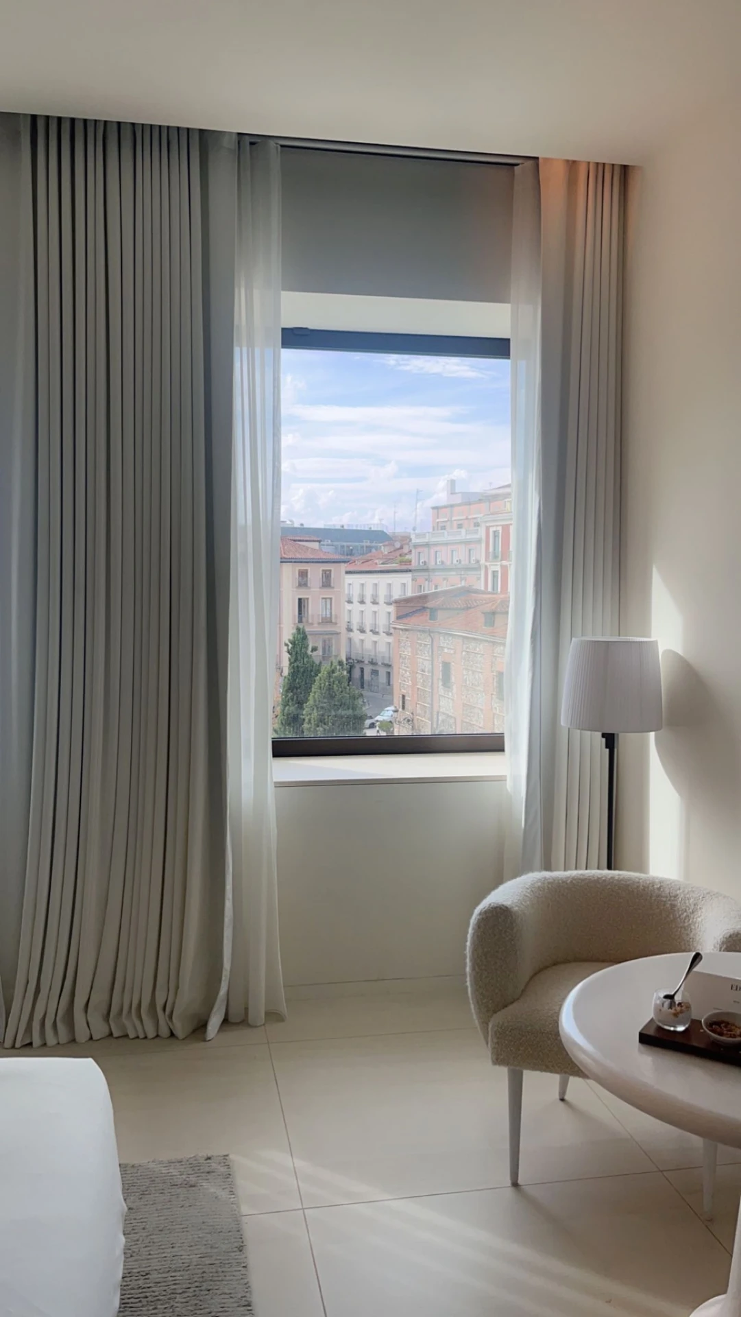 Elegant curtain ideas for the living room with layered curtains combining sheer white drapes and heavier fabric panels, framing a large window with a city view. The space is styled with a cozy boucle armchair, a round white table, and a modern floor lamp, creating a versatile and refined atmosphere filled with natural light.