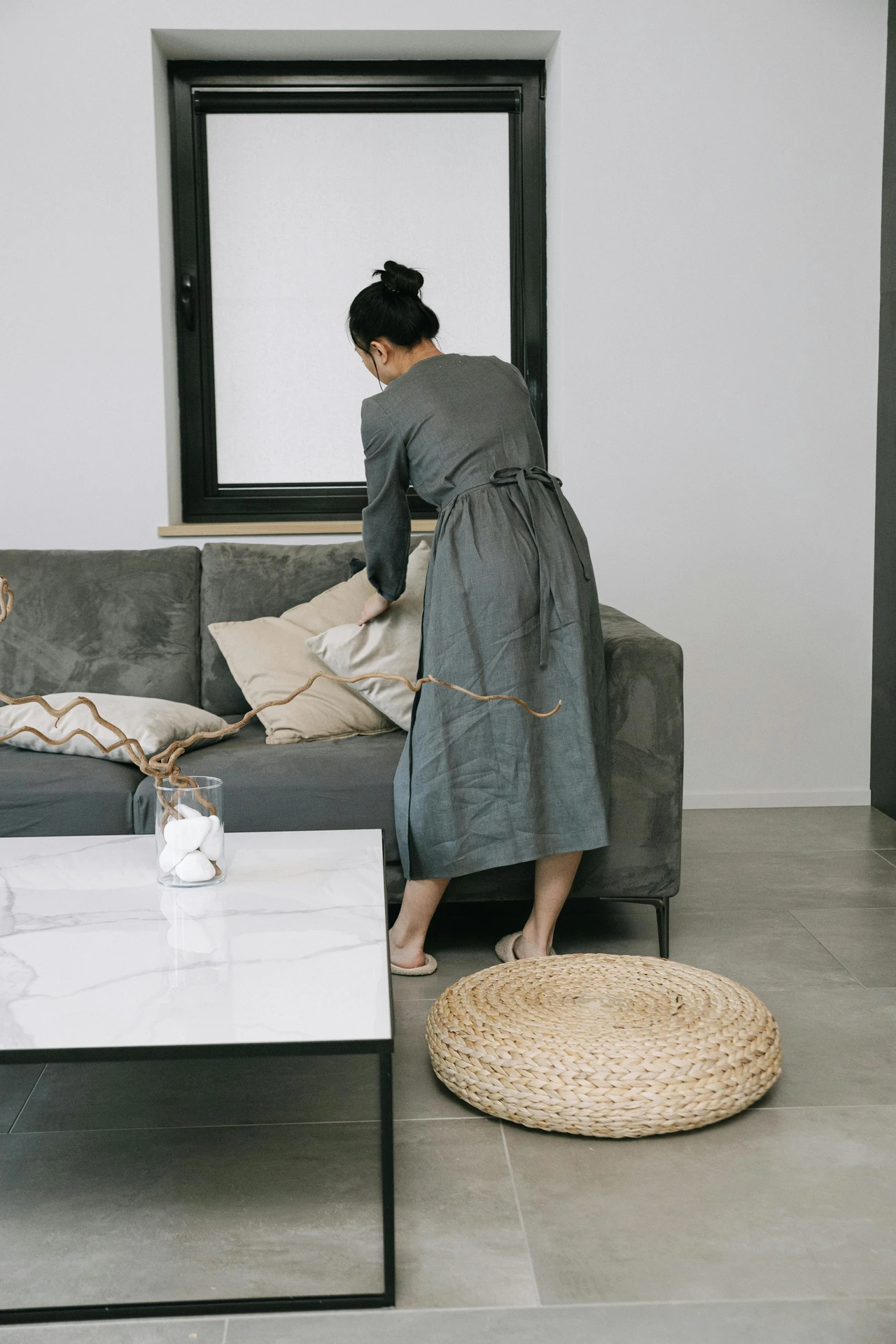 Modern living room with a plush grey sofa styled with soft neutral cushions in beige highlights, adding warmth and contrast to the cool base tone. A marble-top coffee table with minimalist decor and a woven pouf on the floor enhance the natural, calming atmosphere. The setup shows how to style a grey sofa with subtle earthy accents for a balanced and inviting look.