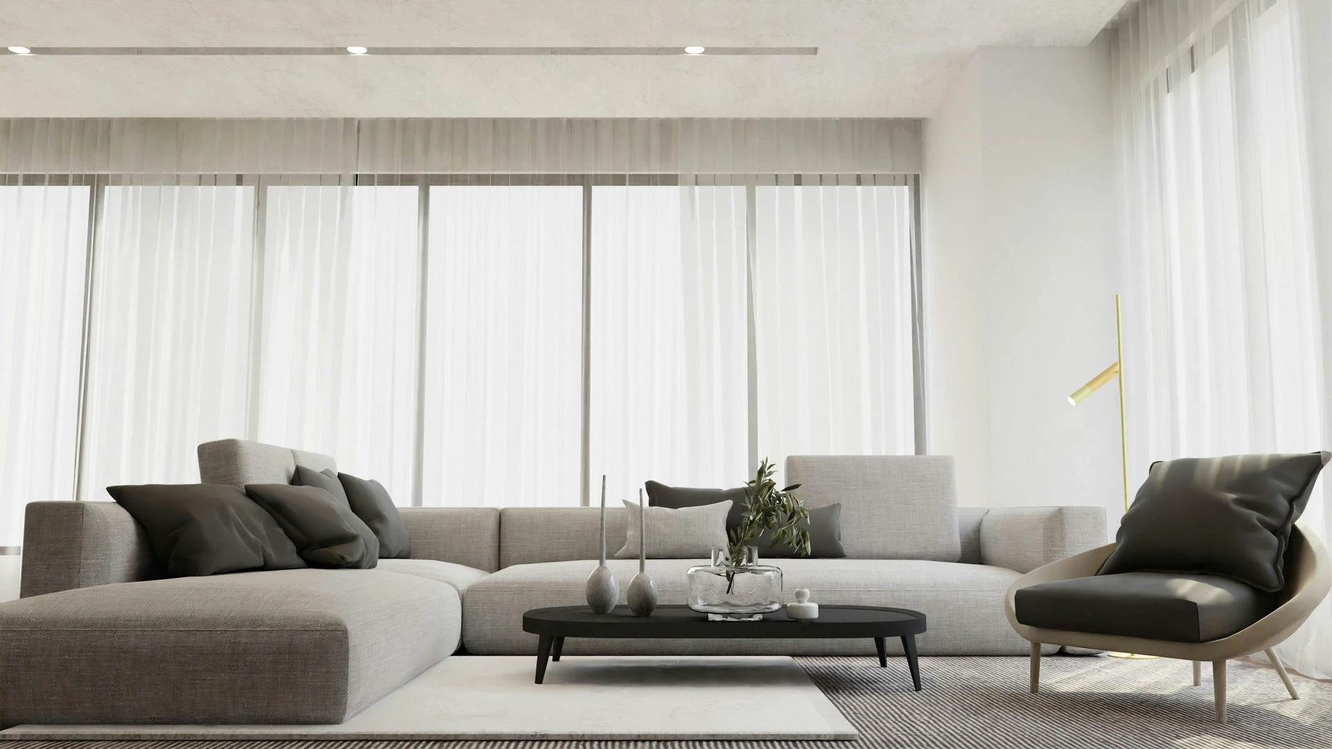 Contemporary living room with a spacious grey sofa styled with sleek black details, including dark throw pillows, a low black coffee table, and a modern lounge chair. Floor-to-ceiling sheer curtains and a gold floor lamp add lightness and elegance to the neutral palette. The setup demonstrates how to style a grey sofa by incorporating bold black accents for contrast, creating a clean yet dynamic modern aesthetic.