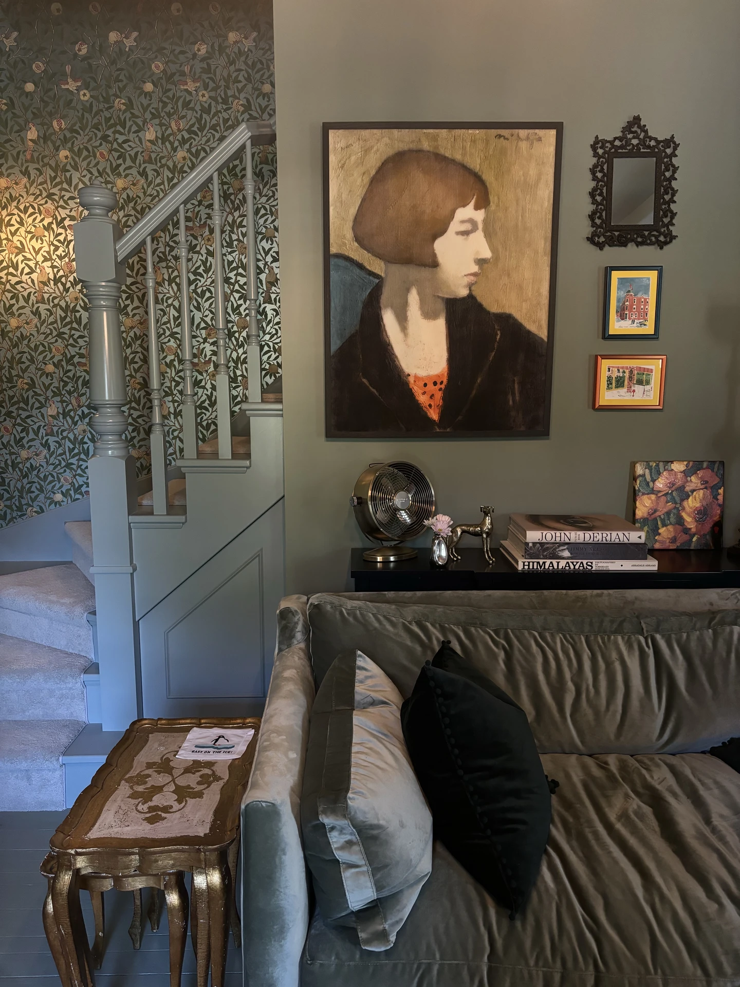 Elegant living room featuring a velvet grey sofa accented with black cushions, paired with an ornate golden side table. Artworks, books, and decorative objects create a curated atmosphere, while wallpaper and framed portraits add personality. The setup illustrates how to style a grey sofa with luxurious details, mixing rich textures, vintage pieces, and layered décor for a sophisticated, character-filled look.
