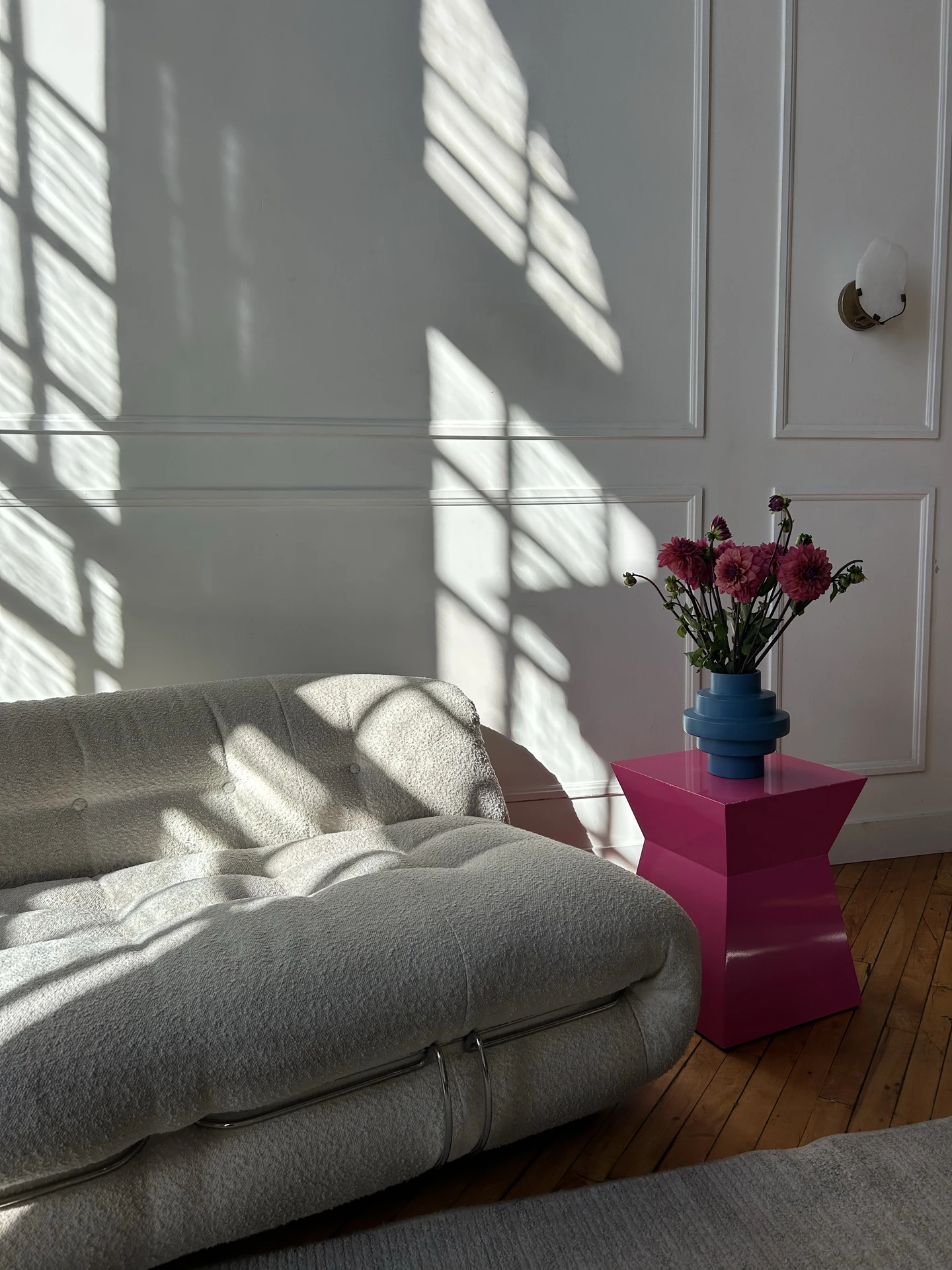 Chic living room with a tufted grey sofa placed against a paneled white wall, bathed in dramatic sunlight and shadows. A bold magenta color pop side table with a sculptural blue vase and pink flowers adds contrast and energy to the neutral backdrop. The look shows how to style a grey sofa by pairing it with vibrant accent furniture that instantly refreshes the space.
