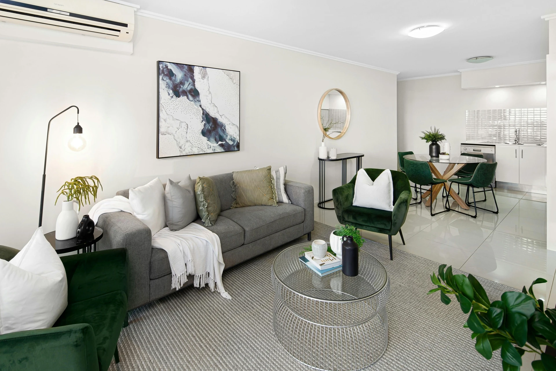 Modern open-plan living room featuring a sleek grey sofa styled with layered cushions and a white throw, paired with two emerald green cozy velvet armchairs. A round glass coffee table with decor, abstract wall art, and plenty of plants add freshness to the space, while the dining area in the background continues the green accents. The look shows how to style a grey sofa by combining it with jewel-toned seating for a balanced yet inviting atmosphere.