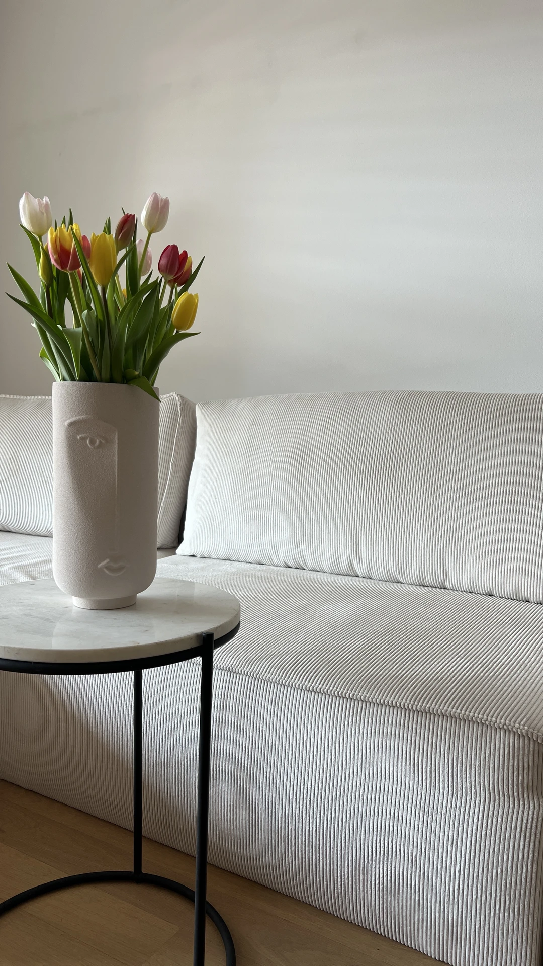 Minimalist living room featuring a sleek grey sofa with subtle pinstripe texture, paired with a round marble side table. A sculptural vase filled with colorful fresh flowers-red, yellow, and pink tulips-adds vibrancy and softness to the neutral space. The setup highlights how to style a grey sofa by bringing in natural elements that create contrast and a lively, welcoming atmosphere.