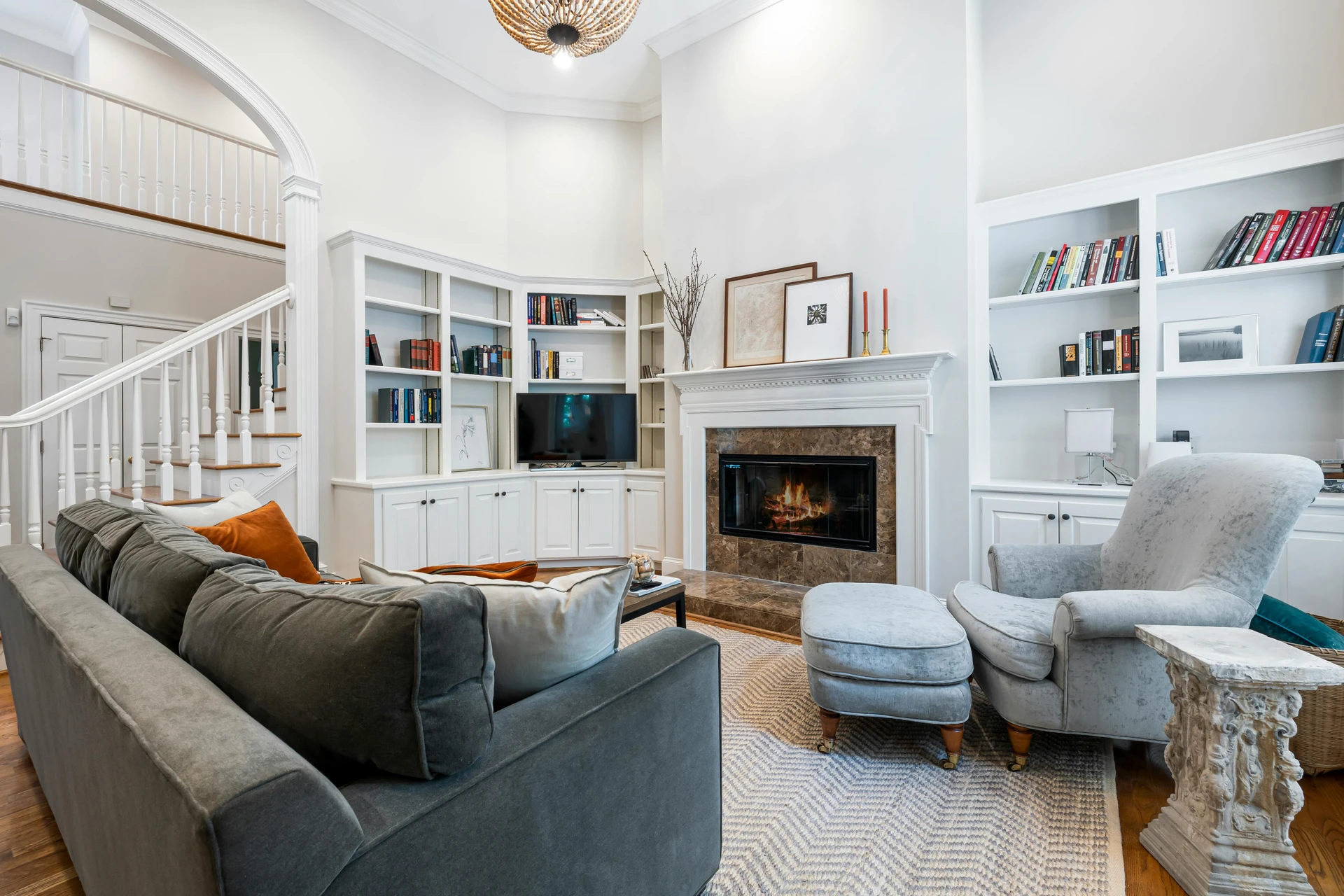Traditional living room with built-in bookshelves, a cozy fireplace, and a grey sofa with grey arm chair arranged around a soft patterned rug. The sofa is styled with warm accent pillows in earthy tones, while the armchair with matching ottoman adds comfort and balance to the space. The setup shows how to style a grey sofa by pairing it with complementary seating in similar tones, creating a cohesive and inviting atmosphere.