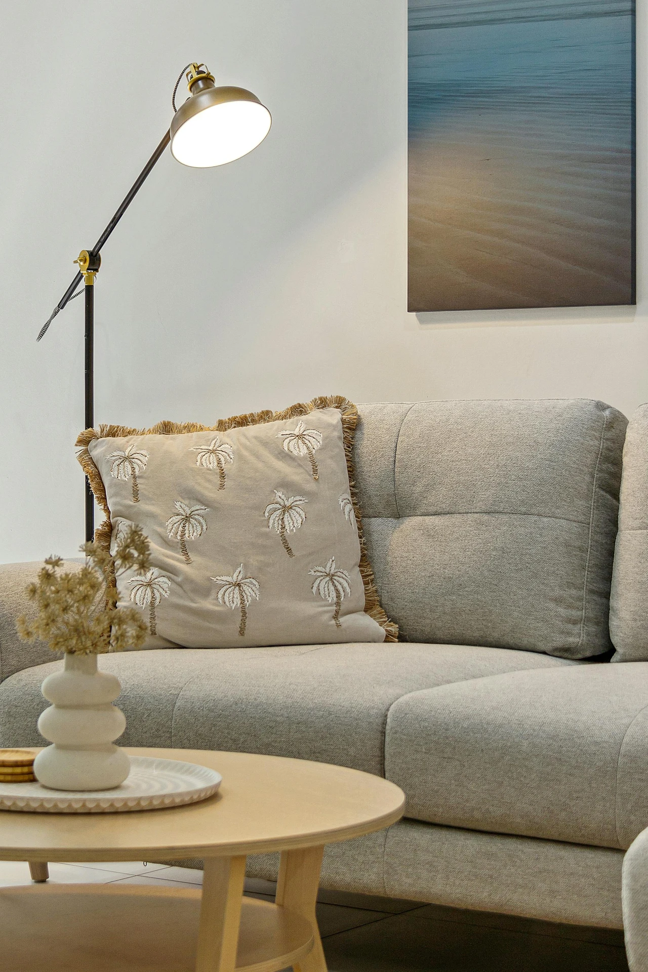 Cozy living room corner with a soft grey sofa styled in boho style, featuring a beige pillow with embroidered palm tree patterns and fringed edges. A round wooden coffee table with a ceramic vase and dried flowers, along with a wall-mounted beach artwork and modern floor lamp, complete the relaxed aesthetic. The setup highlights how to style a grey sofa with natural textures and bohemian accents to create a warm, laid-back atmosphere.