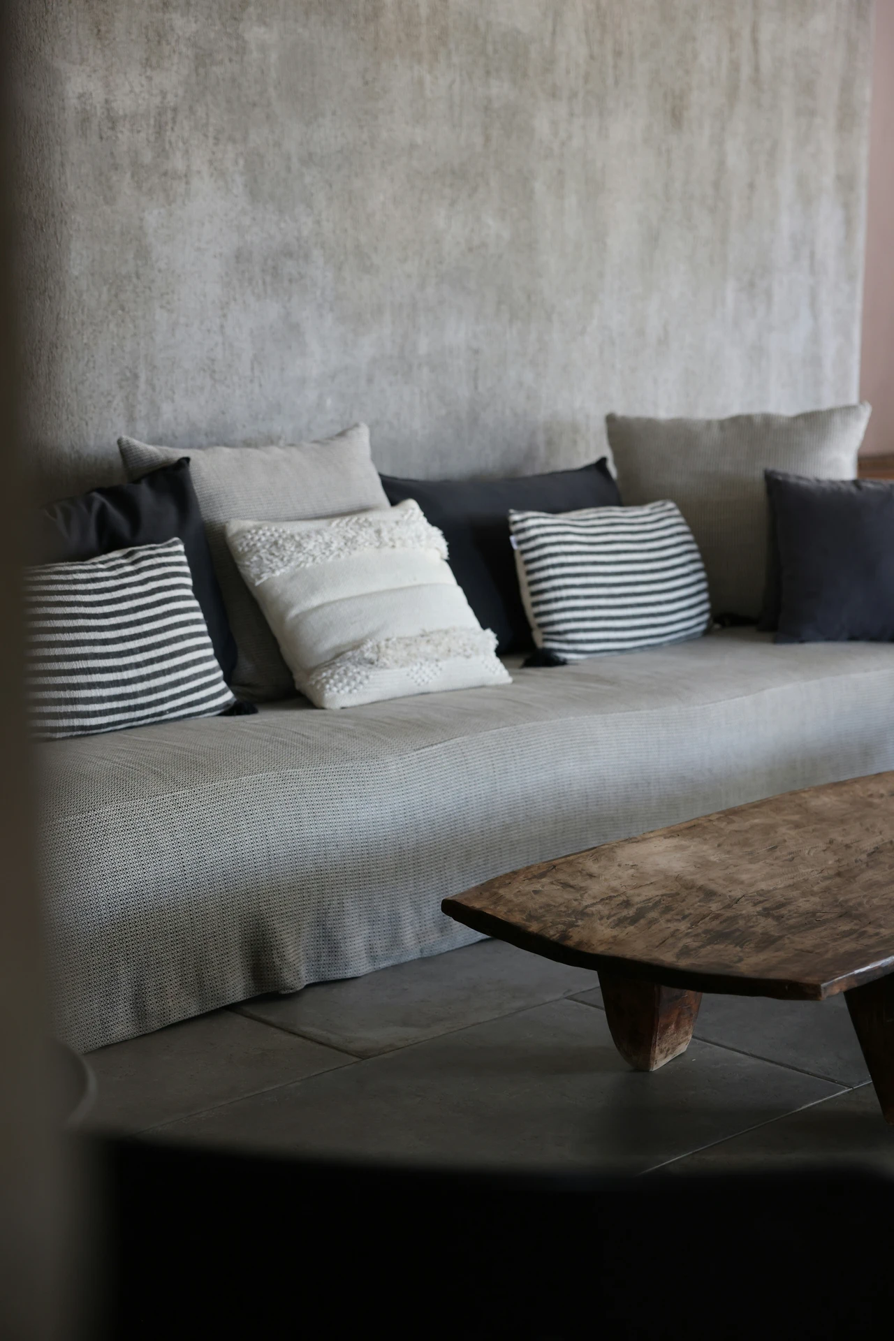 Contemporary living room with a long grey sofa set against a textured concrete wall, styled with a pillow mix of striped, solid, and textured cushions in shades of grey, black, and white. A rustic wooden coffee table sits in front, adding warmth and contrast to the cool-toned palette. The setup highlights how to style a grey sofa by layering different pillow patterns and fabrics for depth and character.