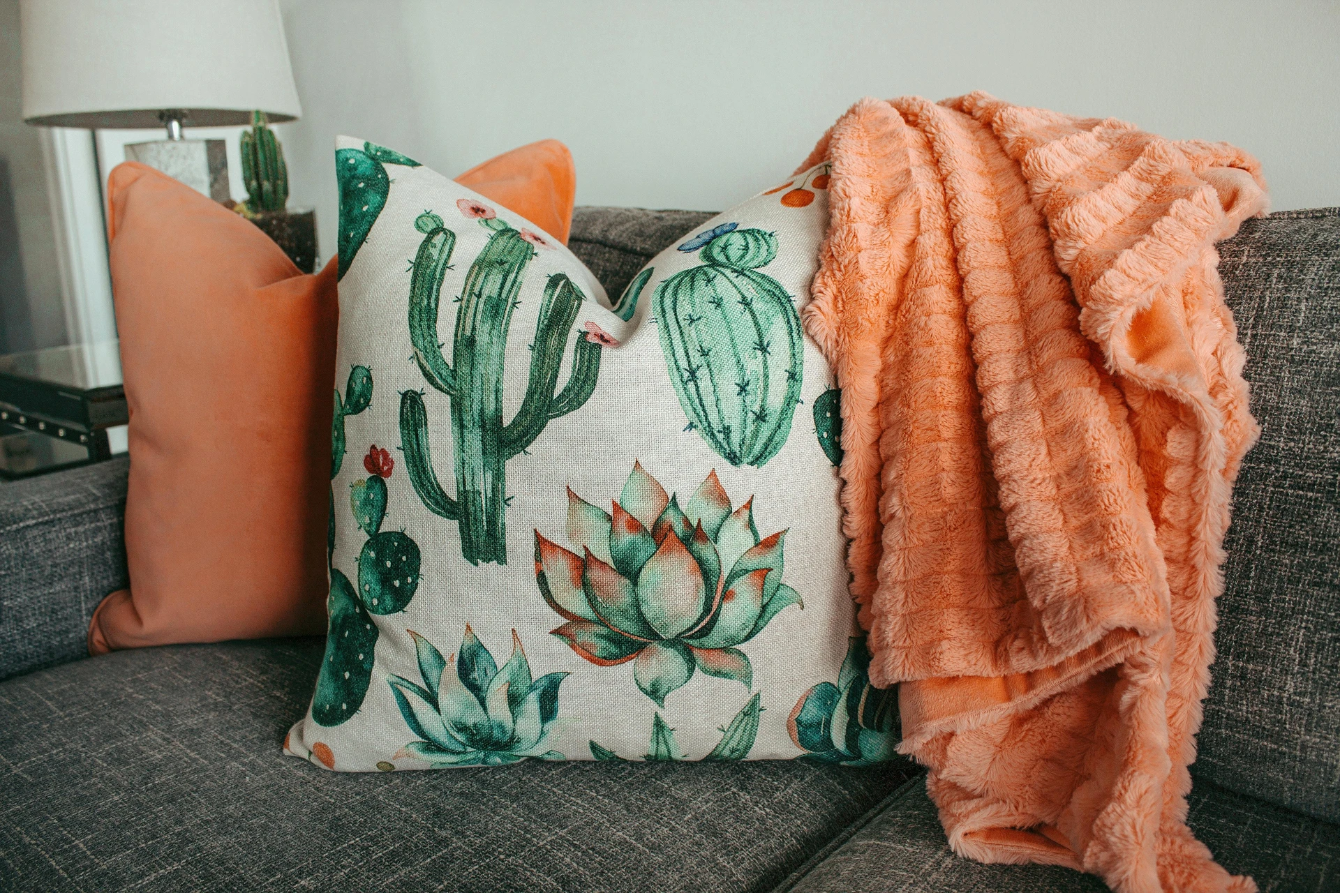 Grey sofa styled with a bold succulents pattern and color pop, featuring a cactus-print pillow, a soft peach throw blanket, and a matching plain orange cushion. The mix of earthy greens and vibrant coral tones adds warmth and character to the neutral base. This setup is a versatile example of how to style a grey sofa, showing how playful patterns and pops of color can instantly brighten and personalize a living room.