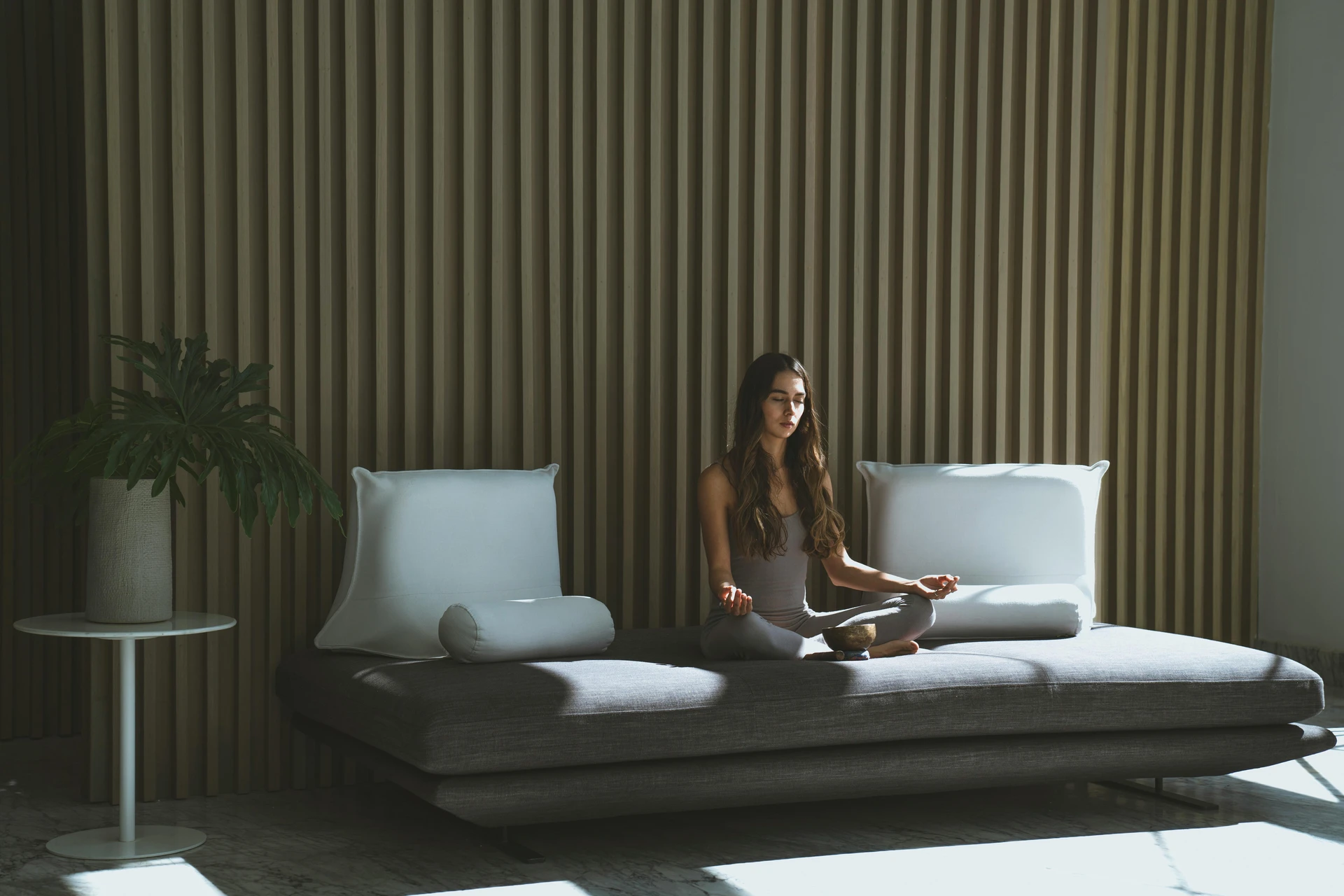 Minimalist living room with a sleek grey sofa styled with large white pillows and cylindrical bolsters, creating a calm and balanced look. A woman sits cross-legged on the sofa meditating, with natural light streaming in through the space and a potted plant adding a touch of greenery. This is an example of how to style a grey sofa, showing how crisp white accents can enhance a serene, modern, and uncluttered aesthetic.