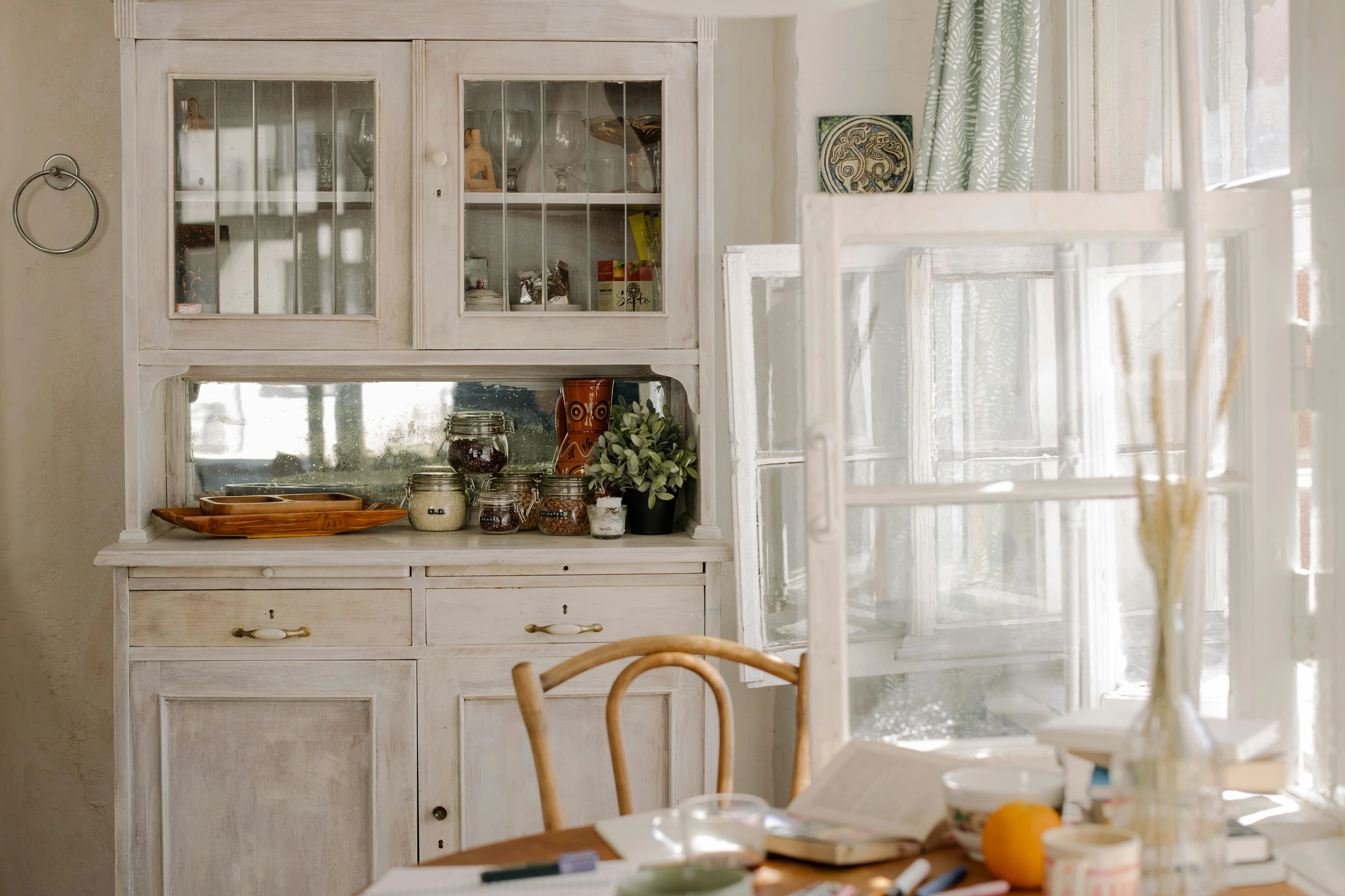 Bright vintage-style kitchen setup demonstrating smart kitchen counter organization with the idea to add an additional kitchen cabinet. A distressed white wooden hutch with glass doors holds glassware on top, while the counter space below features neatly arranged glass jars filled with dry goods, a wooden tray, a decorative mug, and a small potted plant. Sunlight streams in through open windows, creating a warm and inviting atmosphere.
