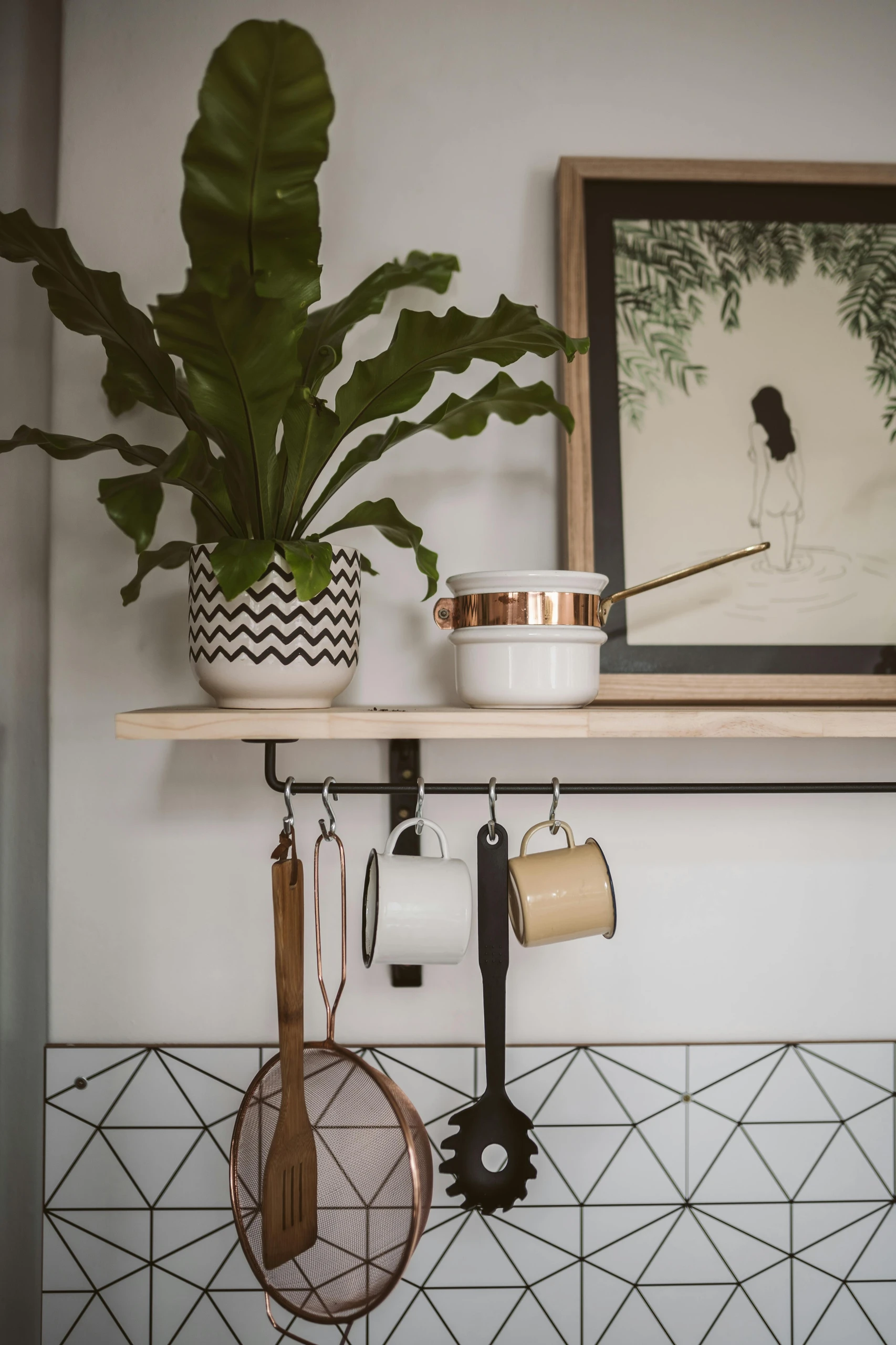 A stylish kitchen counter organization idea showing how to attach hooks under your floating shelf for added storage. A light wooden floating shelf holds a potted plant in a chevron-patterned pot, a framed artwork, and a small white saucepan with a copper accent. Beneath the shelf, black hooks neatly display kitchen essentials like a spatula, strainer, pasta spoon, and two ceramic mugs, keeping them within easy reach while freeing up counter space.