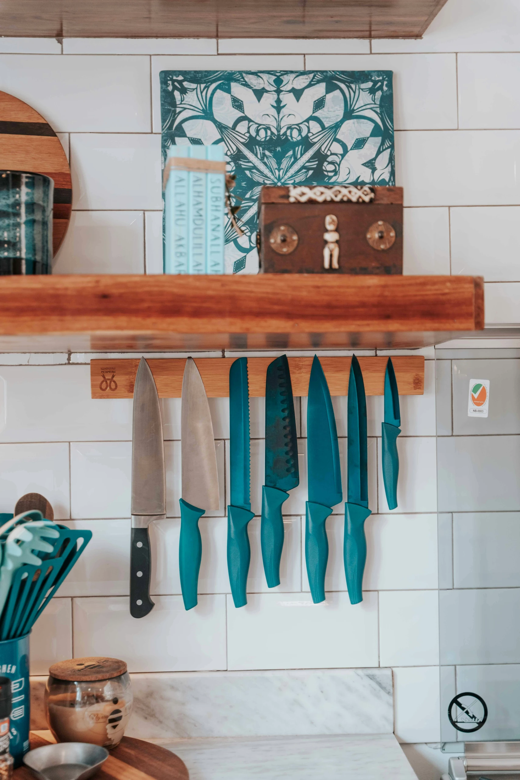 A creative kitchen counter organization idea featuring a set of knives neatly displayed on a magnetic strip mounted to a white subway tile wall. The setup shows how you can attach your knives to magnets on the wall, keeping them easily accessible while freeing up counter and drawer space. Above, a rustic wooden shelf holds decorative items like a small wooden chest, books, and a patterned cutting board, blending practicality with personal style.