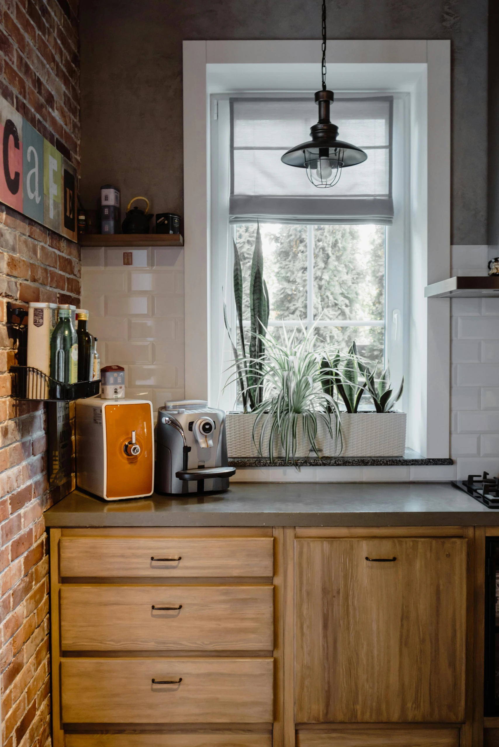 A cozy rustic kitchen setup showcasing clever kitchen counter organization with a dedicated coffee station. An orange water dispenser and a sleek silver espresso machine sit neatly on the countertop beside a small wall-mounted shelf holding bottles. The backdrop features exposed brick, white subway tiles, and a bright window adorned with lush potted plants, blending function and charm in a versatile space.