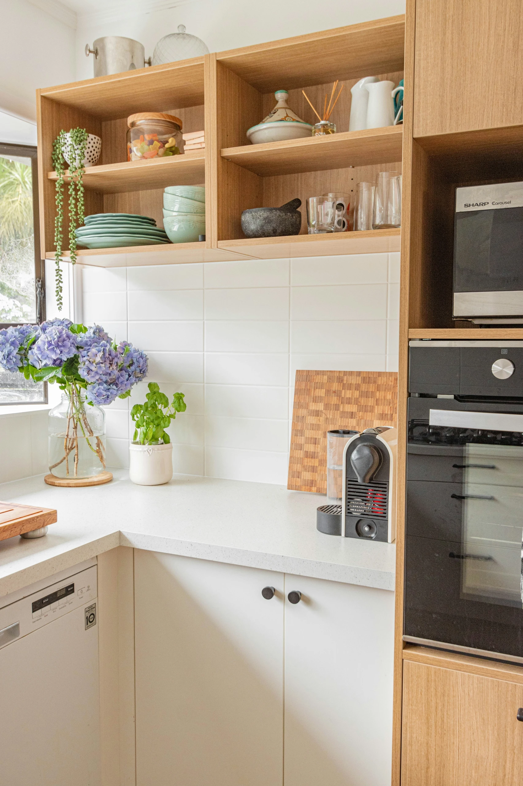 A bright and airy kitchen corner showcasing smart kitchen counter organization, with a clean white countertop holding only a cutting board, a coffee machine, and a small herb plant. Open wooden shelves above display neatly stacked plates, bowls, and decorative items, while a vase of vibrant purple hydrangeas adds a fresh touch by the window. A perfect example of how to declutter your kitchen counter for a functional yet inviting space.