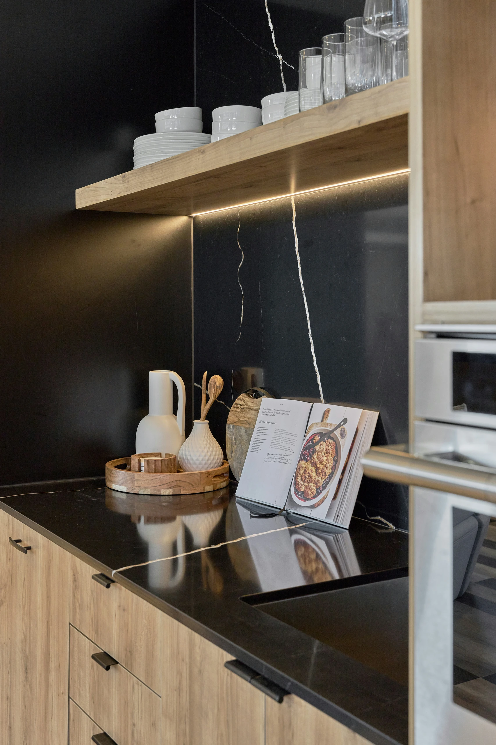 A sleek and modern kitchen counter organization setup with a black countertop, light wood cabinets, and a matching wooden floating shelf holding neatly stacked white plates, bowls, and clear glasses. On the counter, a round wooden tray groups utensils on trays, including a ceramic pitcher, a textured vase with wooden spoons, and small accessories, creating a tidy yet stylish arrangement. An open cookbook rests beside the tray, making the space both functional and visually appealing.